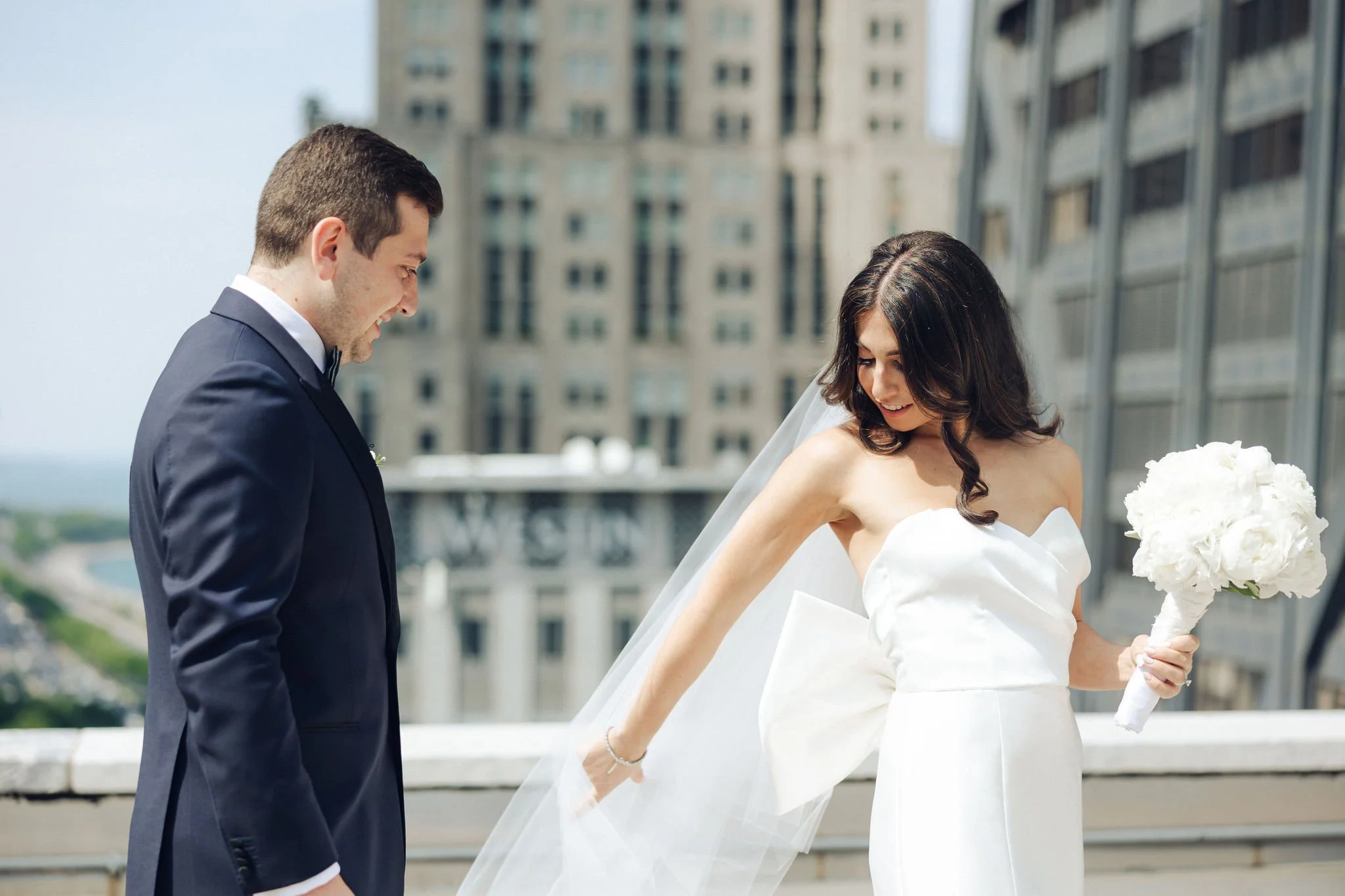 A bride and groom smiling on a rooftop with tall buildings in the background during their wedding. The bride is in a white strapless dress holding a bouquet, and the groom is in a navy blue suit.