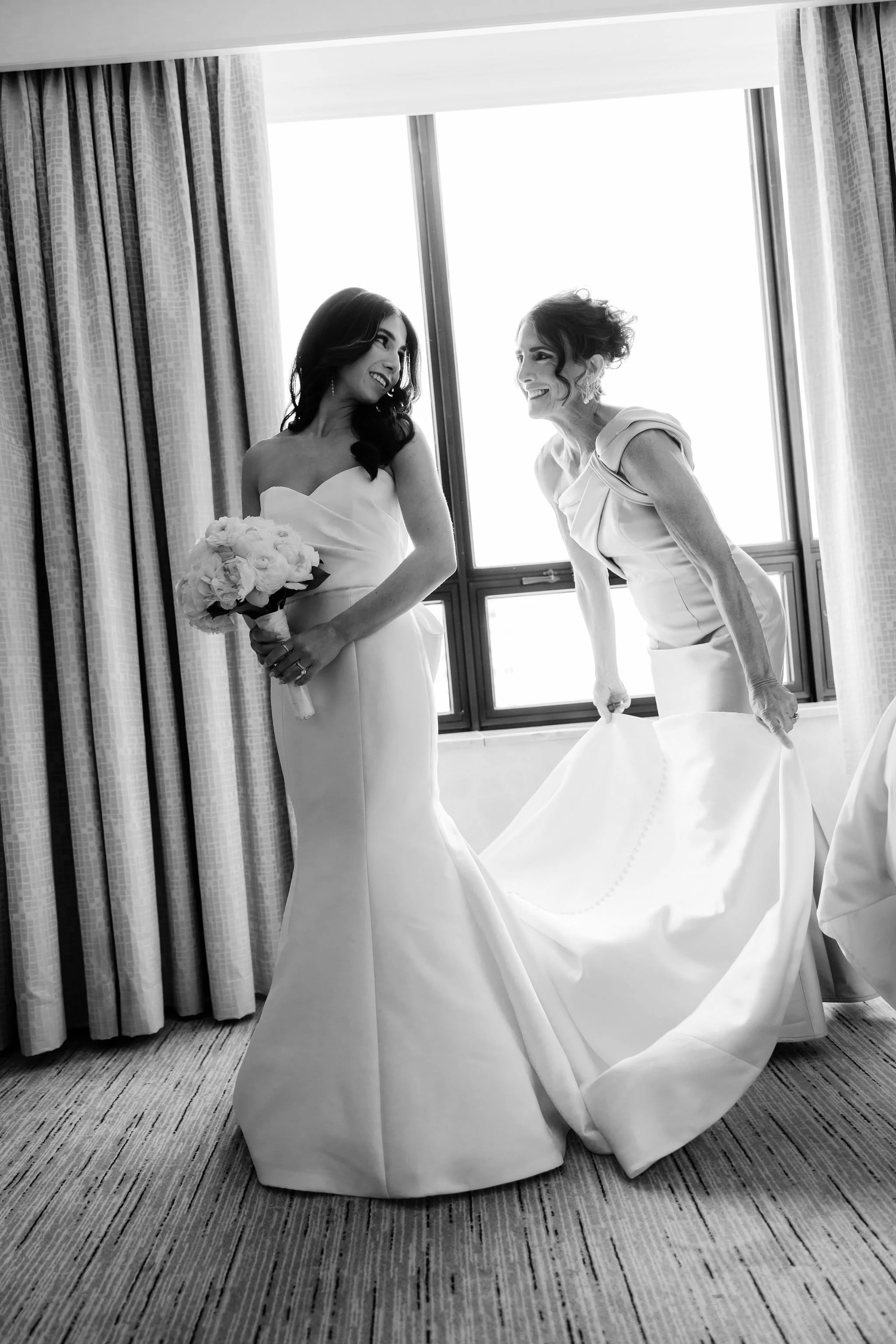 Black and white photo of a bride holding a bouquet of flowers, smiling at another woman in a wedding dress, in front of a window with curtains.