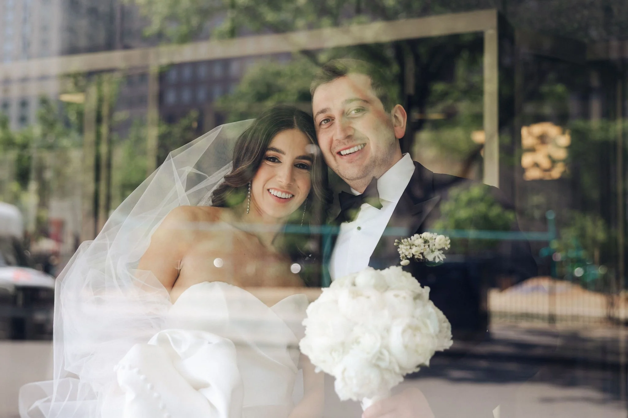 Bride and groom looking through glass window, smiling, with the bride holding a white bouquet, reflections of street and buildings visible in the glass.
