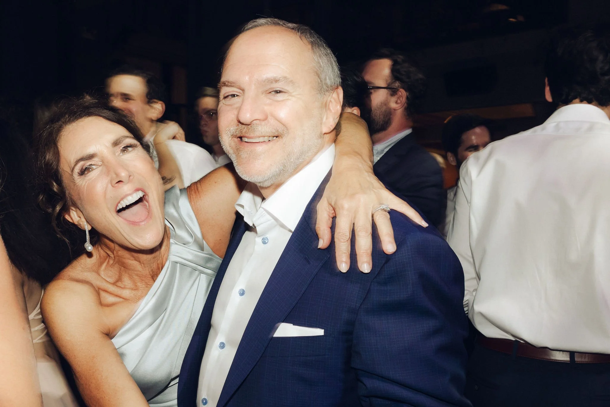 A woman and a man smiling at a social event, the woman has her arm around the man's shoulders.