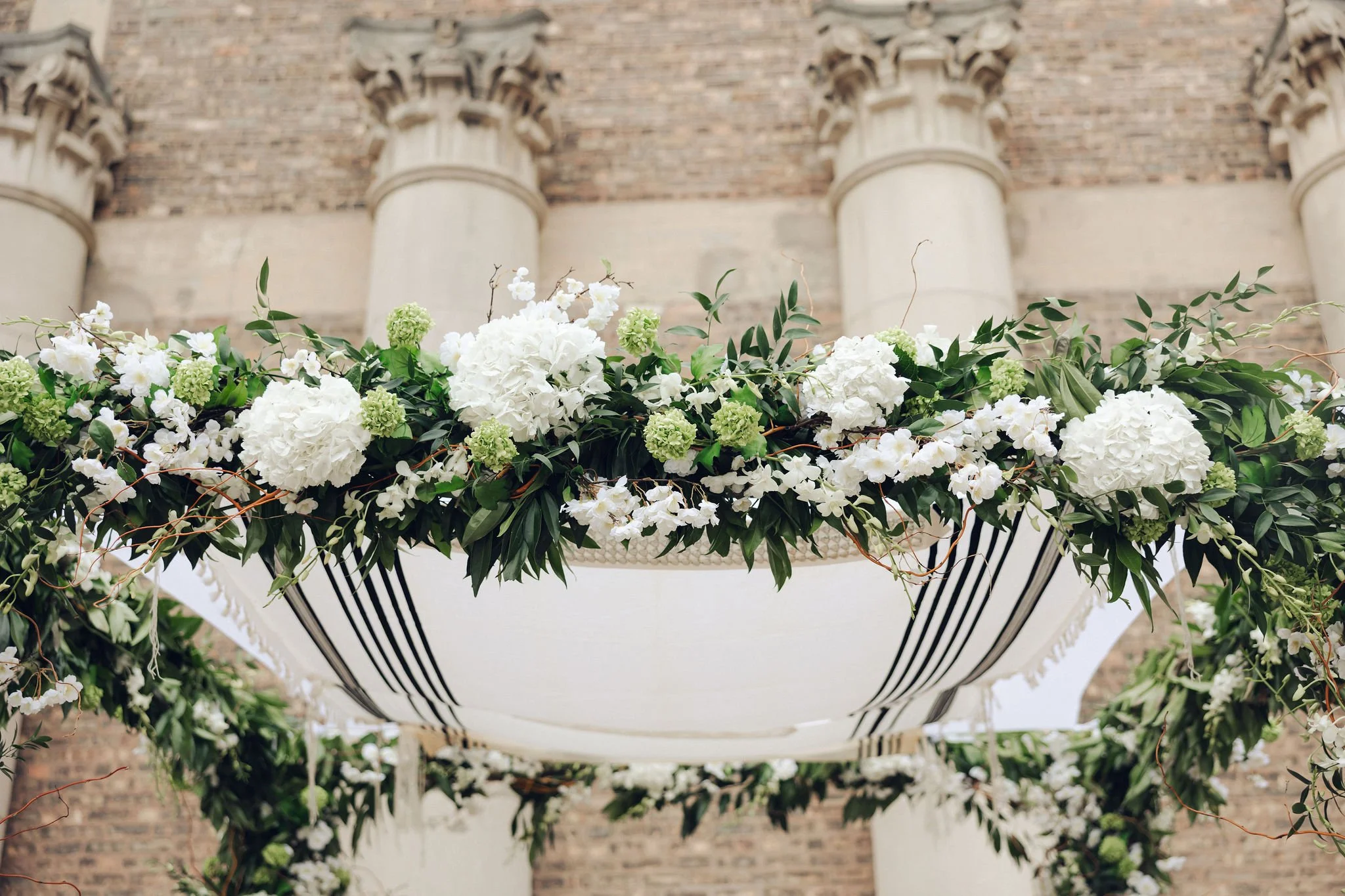 A floral arrangement with white and green flowers and greenery hanging from a ceiling above a brick building.