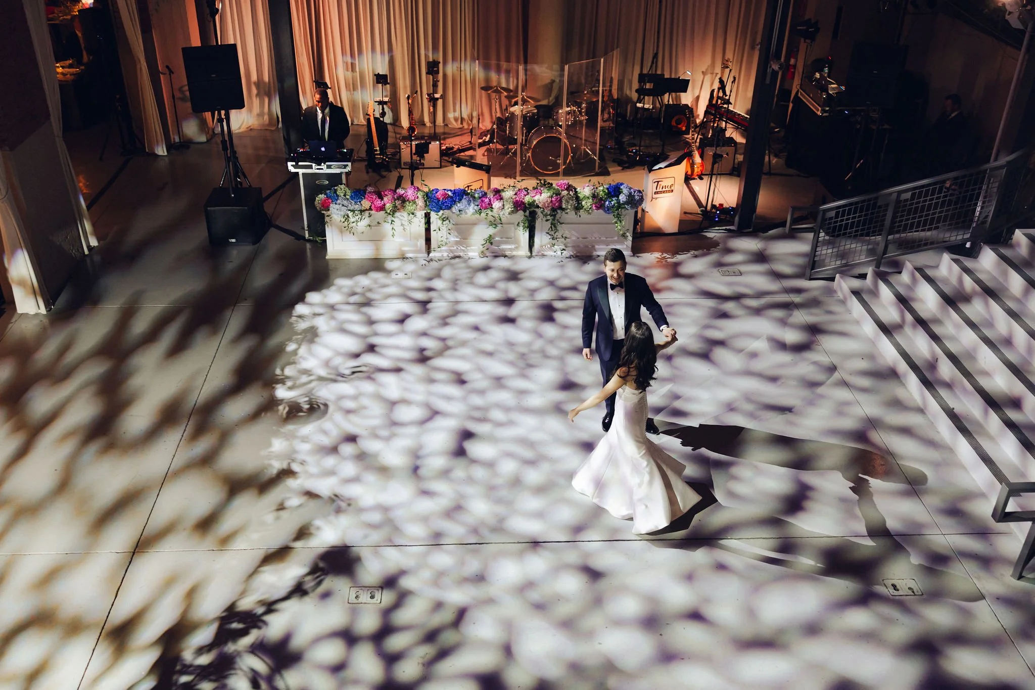 A bride and groom are dancing on a decorated dance floor at their wedding reception, with a live band setup and floral arrangements in the background.