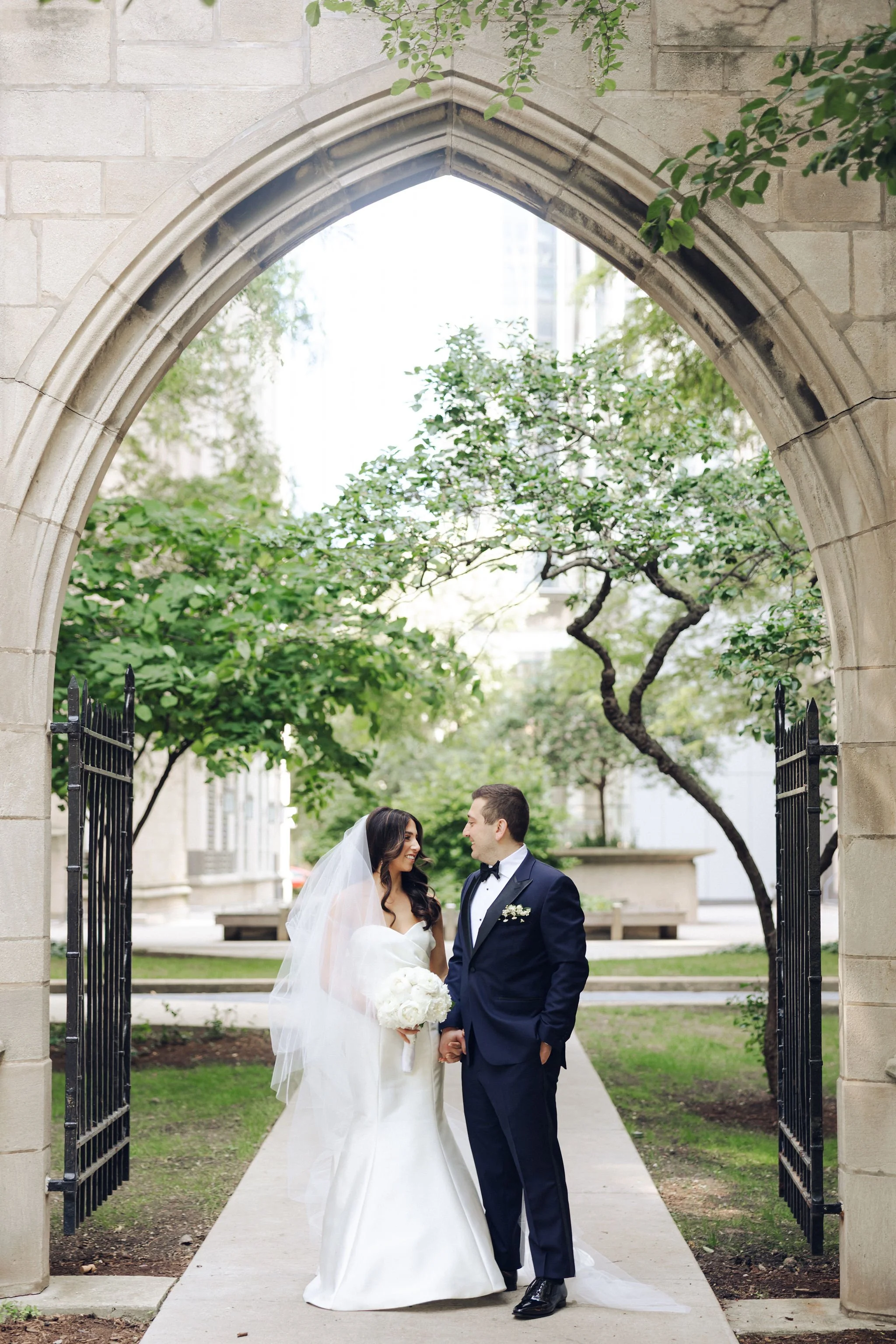 Bride and groom holding hands and smiling under an archway in a garden setting, with trees and a city building in the background.