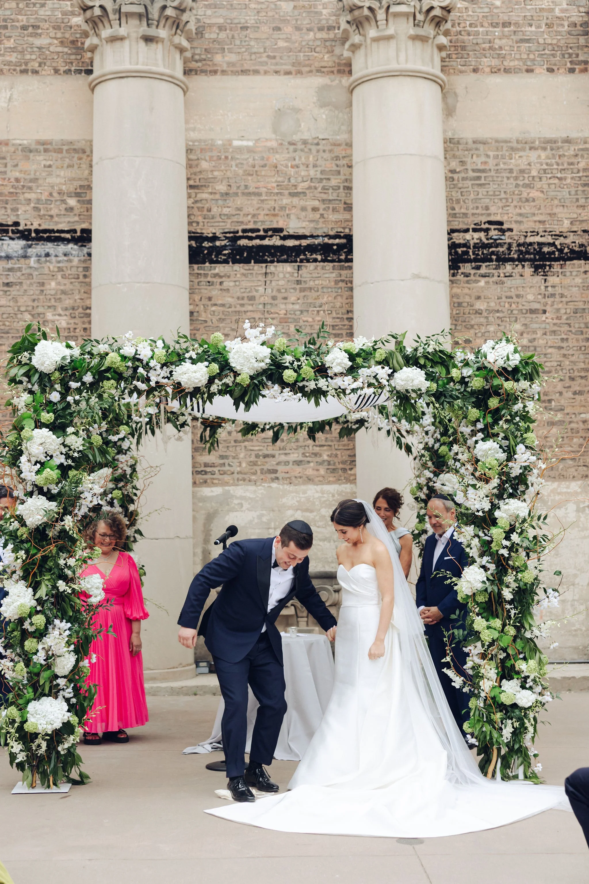 A bride and groom getting married under a floral arch, with ceremony officiants and guests present in an indoor venue with brick walls and columns.