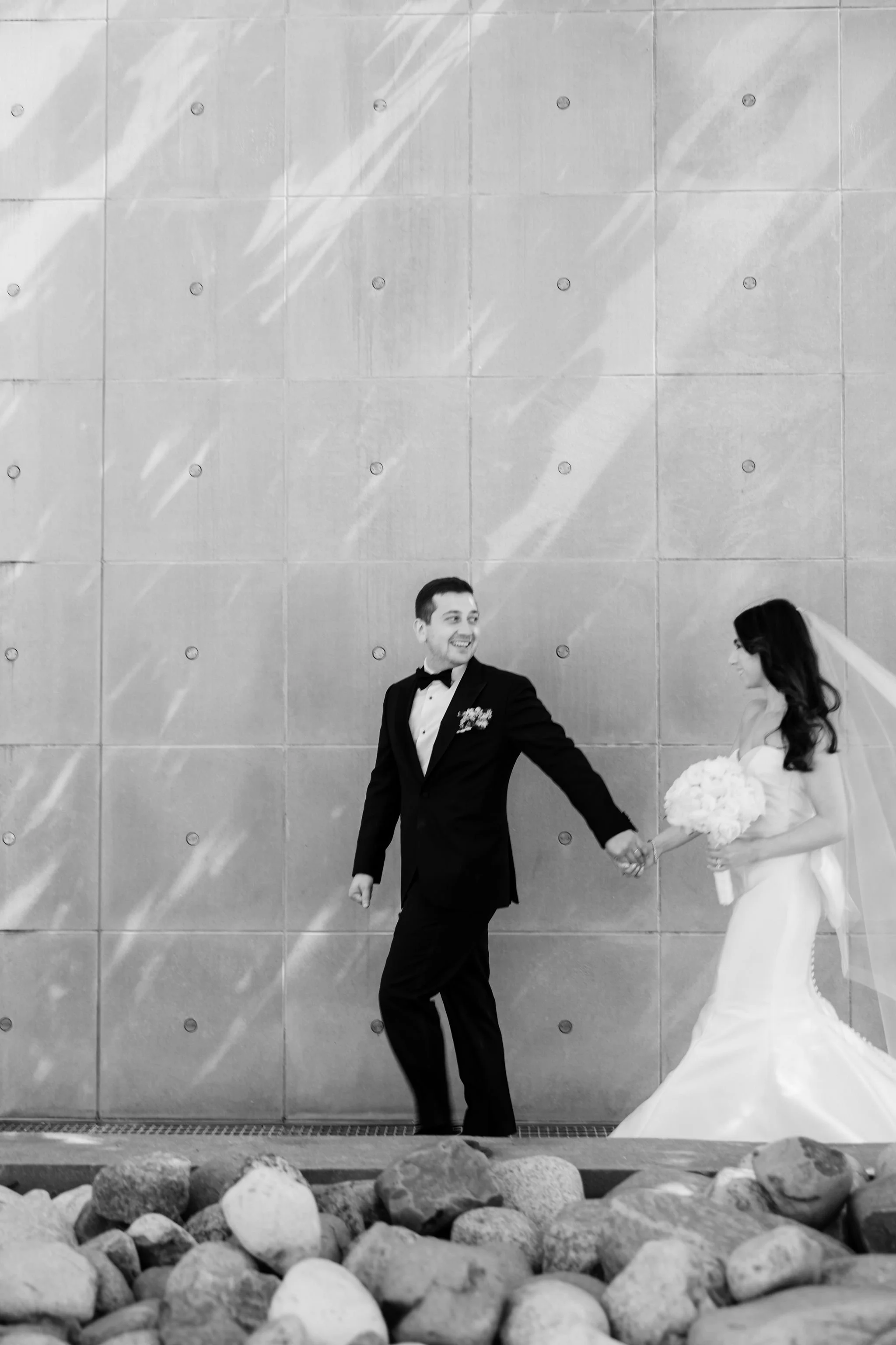 Black and white photo of a bride and groom holding hands and walking together, with the groom smiling and looking at the bride, against a tiled wall background.