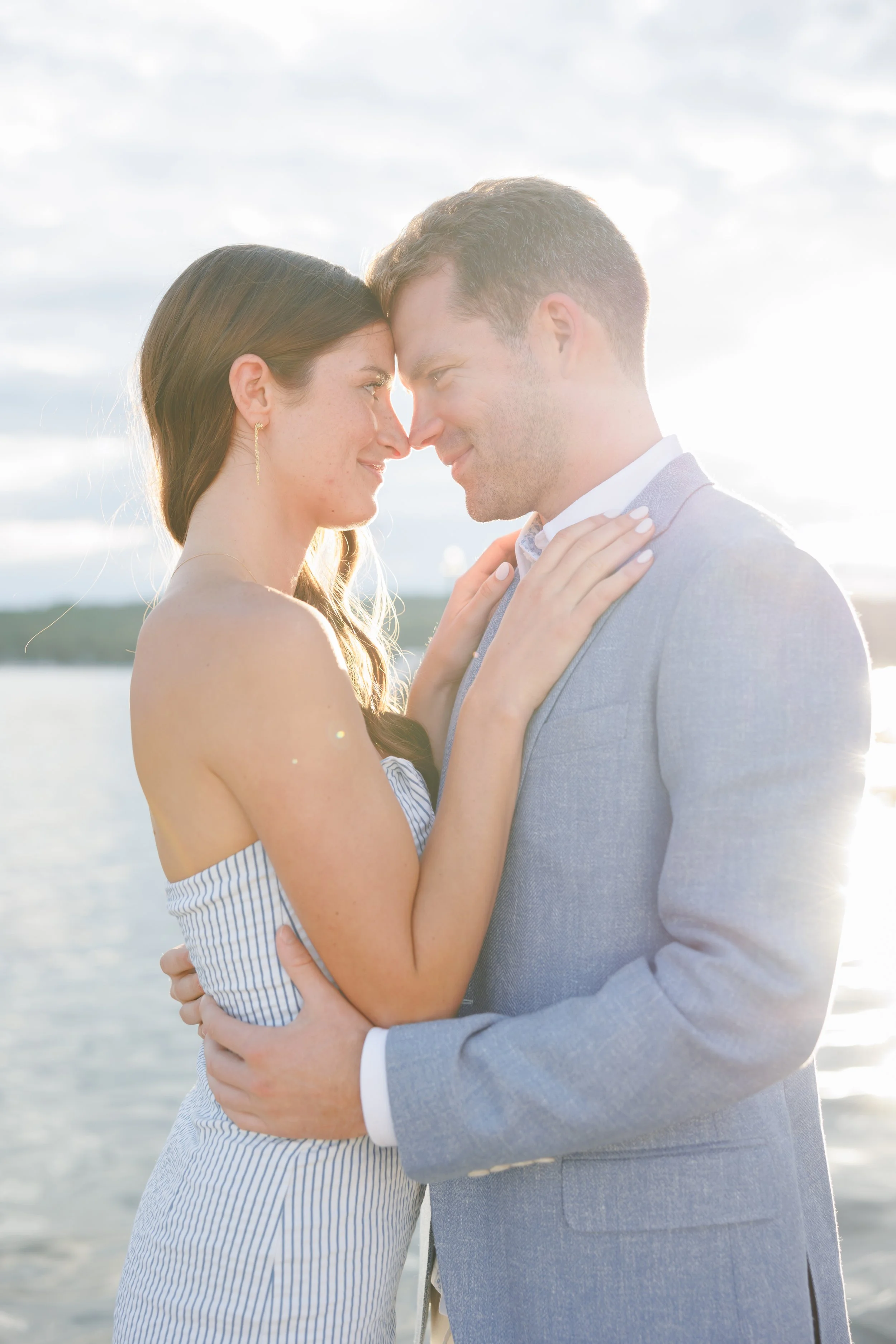 A couple is standing close together outdoors at sunset, with their foreheads touching and smiling at each other, near a body of water.