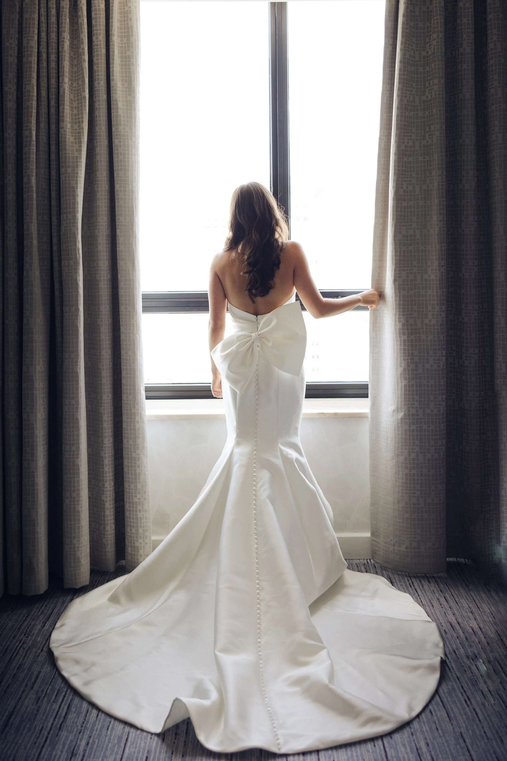 A woman in a strapless wedding gown with a large bow on the back stands by a window, holding a curtain open, looking outside.