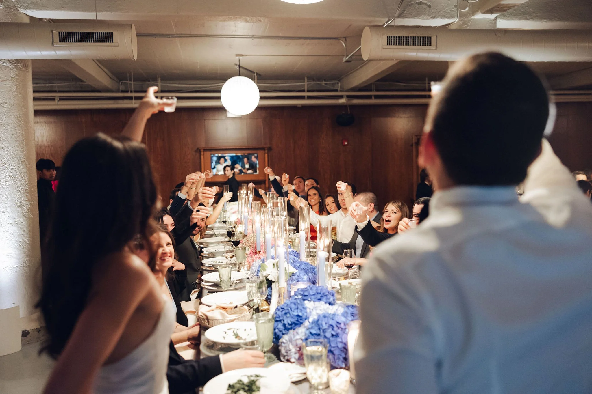 People raising glasses at a wedding reception or banquet dinner, seated at a long decorated table with floral centerpieces and candles.