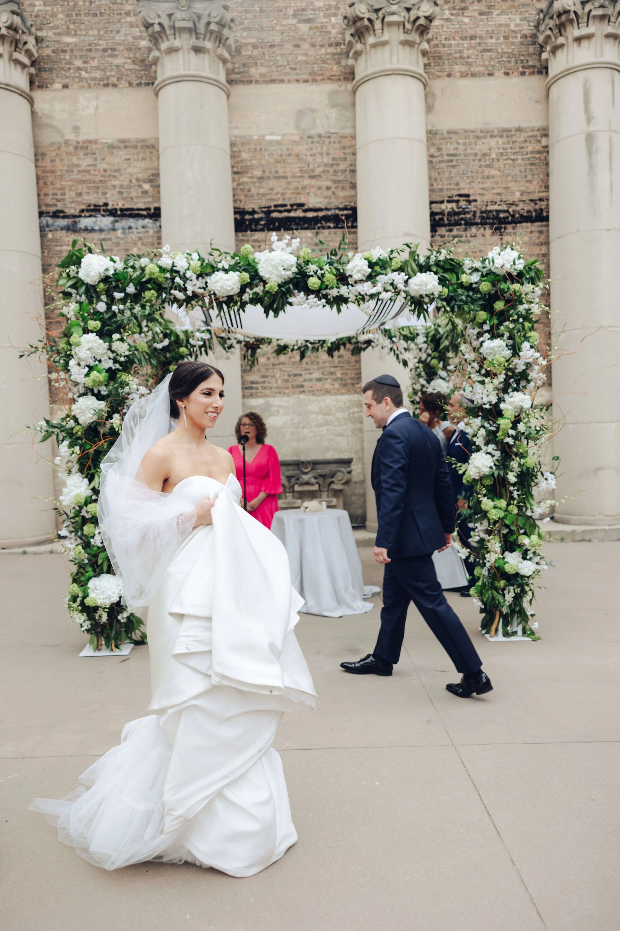 Bride in a white wedding gown during a wedding ceremony outdoors with floral arch and brick columns in the background.