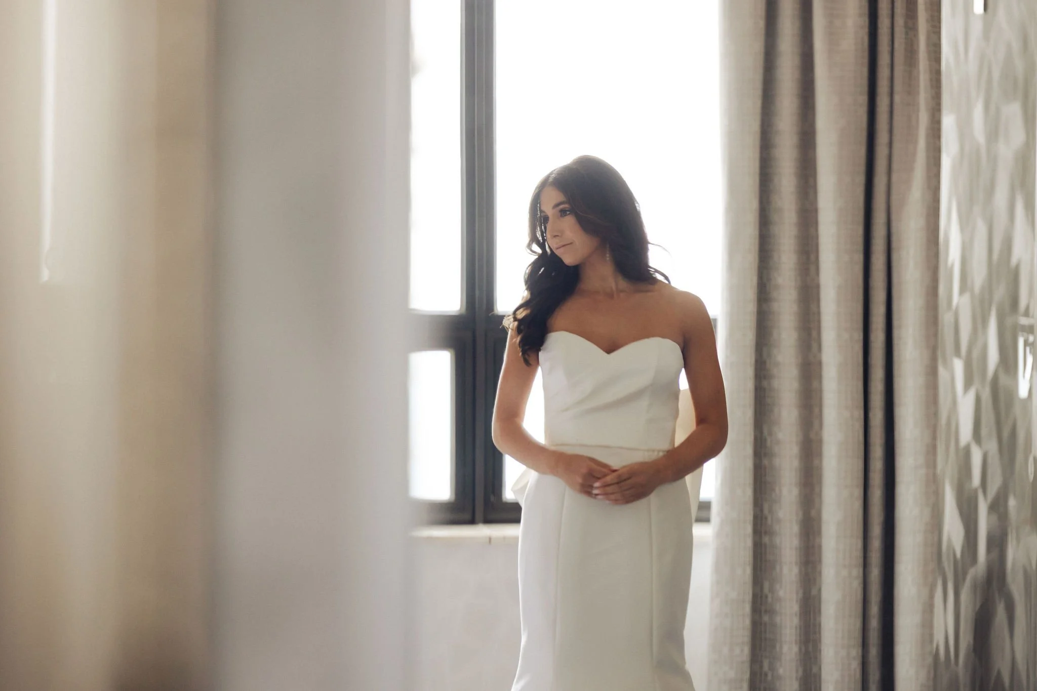 A woman in a strapless white wedding dress standing by a window with curtains, looking to the side in an elegant room with patterned wallpaper.