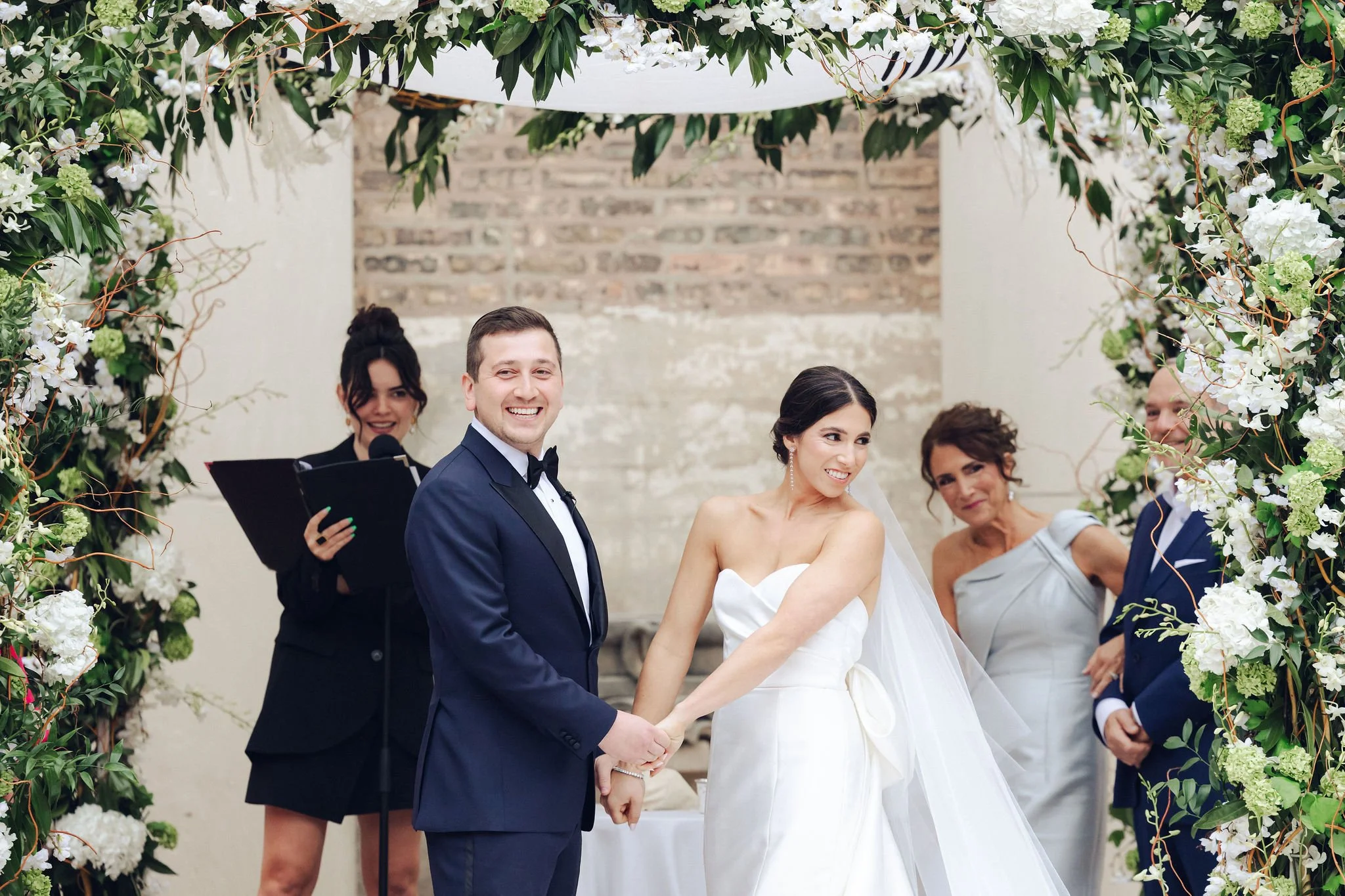 A bride and groom holding hands at their wedding ceremony, smiling. The bride is wearing a white strapless wedding dress and veil, and the groom is in a dark tuxedo. They are surrounded by floral decorations and friends or family members, with one pe