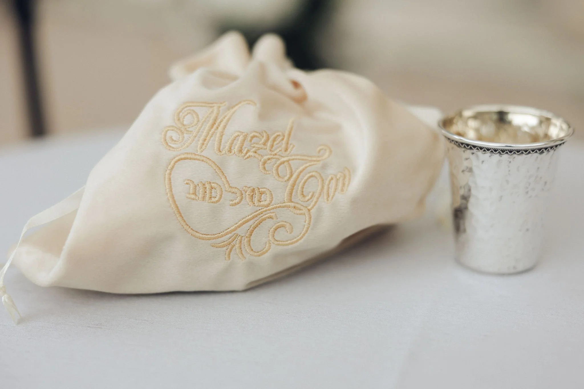 A white satin drawstring bag embroidered with gold thread reading 'Mazel Tov' and a silver chalice on a white tablecloth.