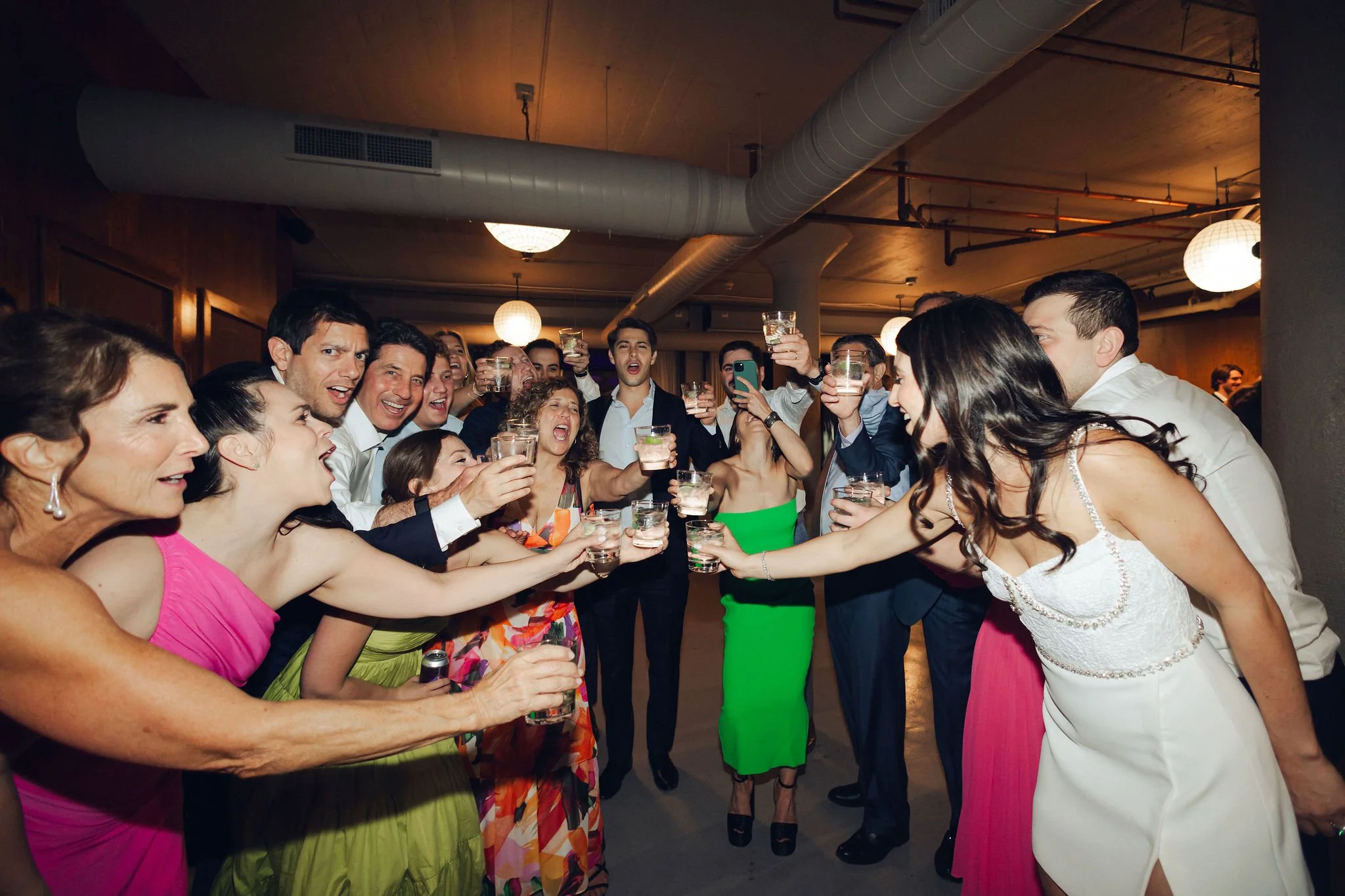 Group of people at a party raising glasses for a toast, some women are dressed in colorful dresses, and some men in suits, in a warmly lit indoor setting.
