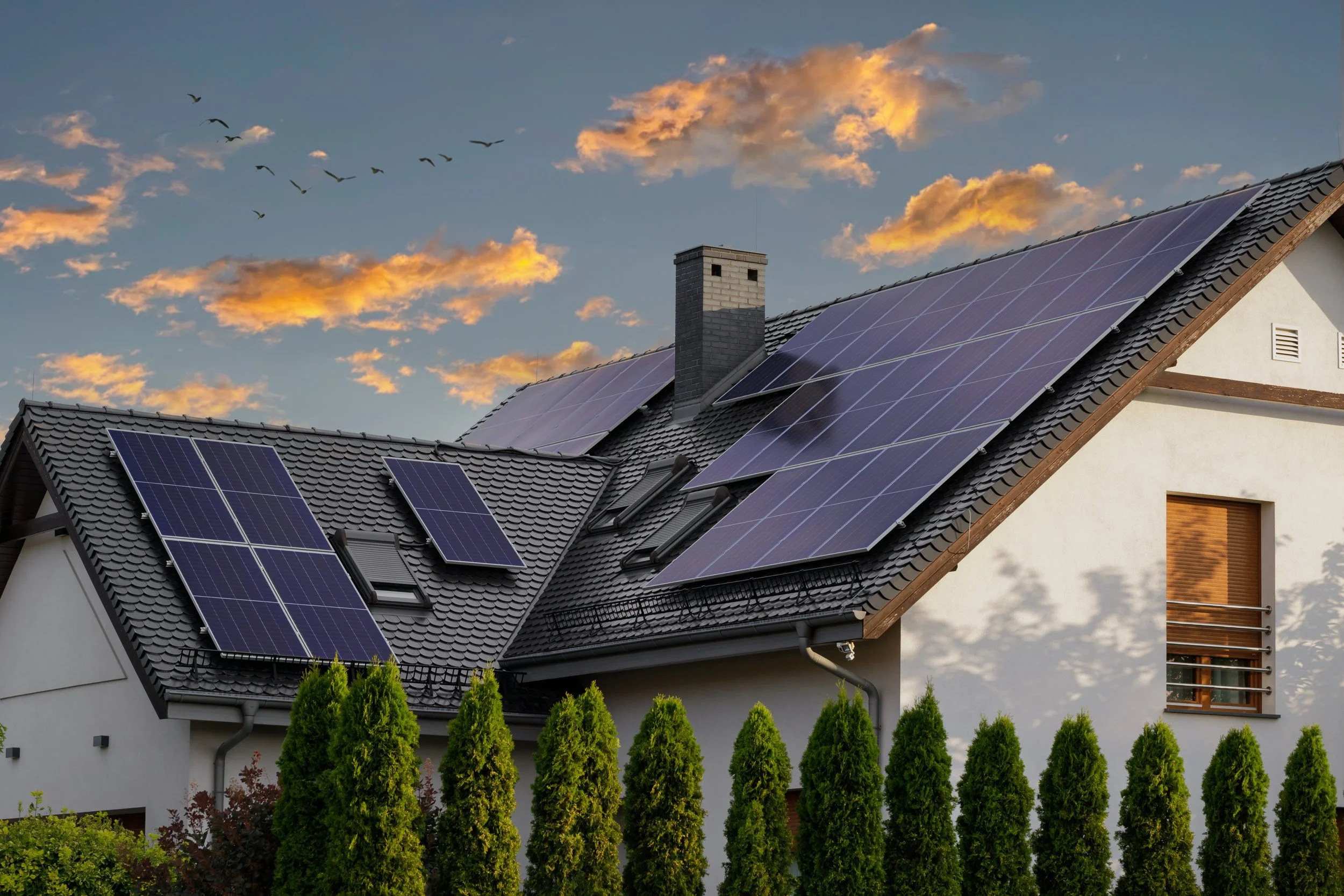 House with solar panels on the roof during sunset, surrounded by green trees.