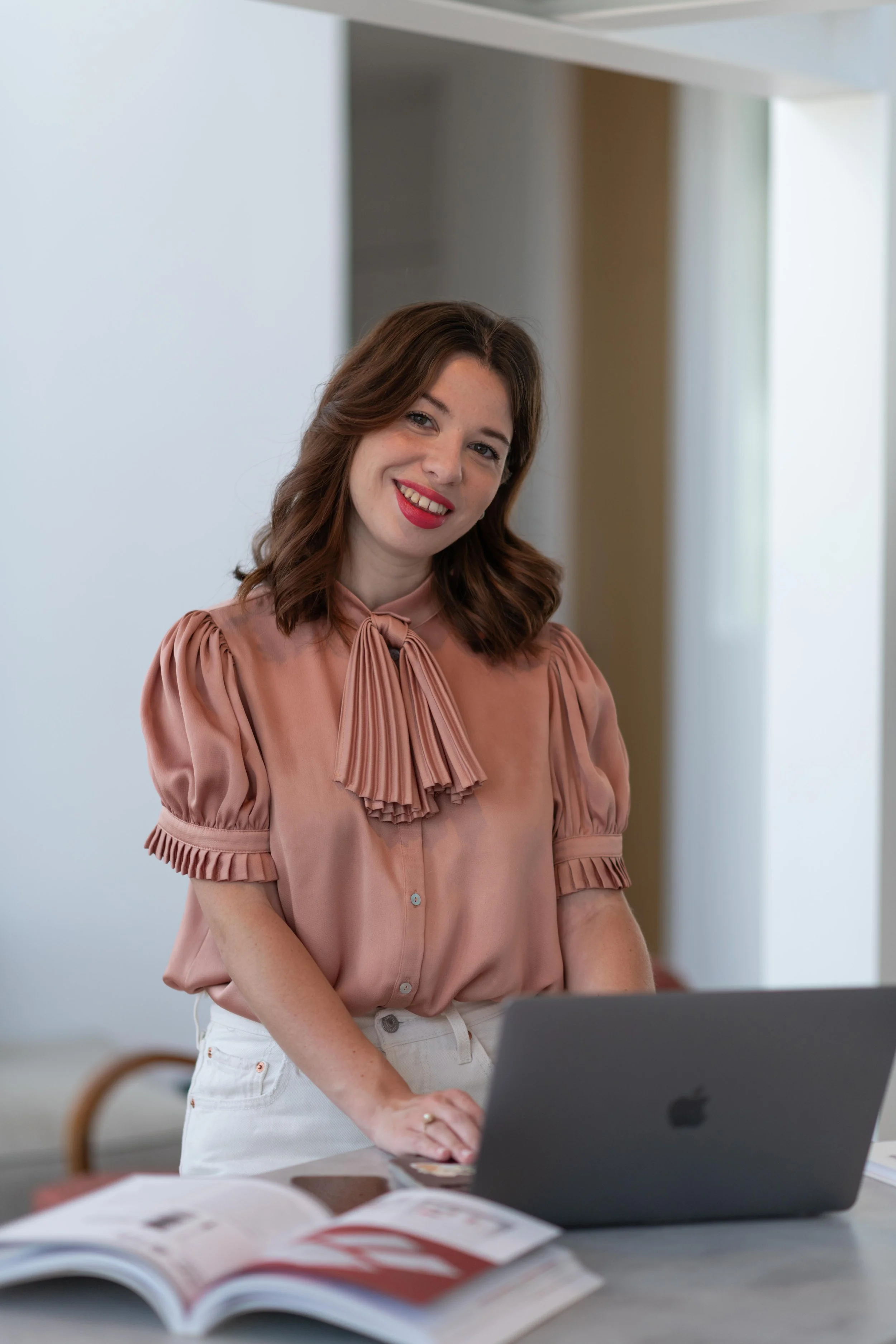Femme brune, Eva LECONTE, du podcast Comme un gant, portant une blouse rose à manches bouffantes avec un nœud à l'encolure, se tient derrière un ordinateur portable sur une table, avec un magazine ouvert devant elle.