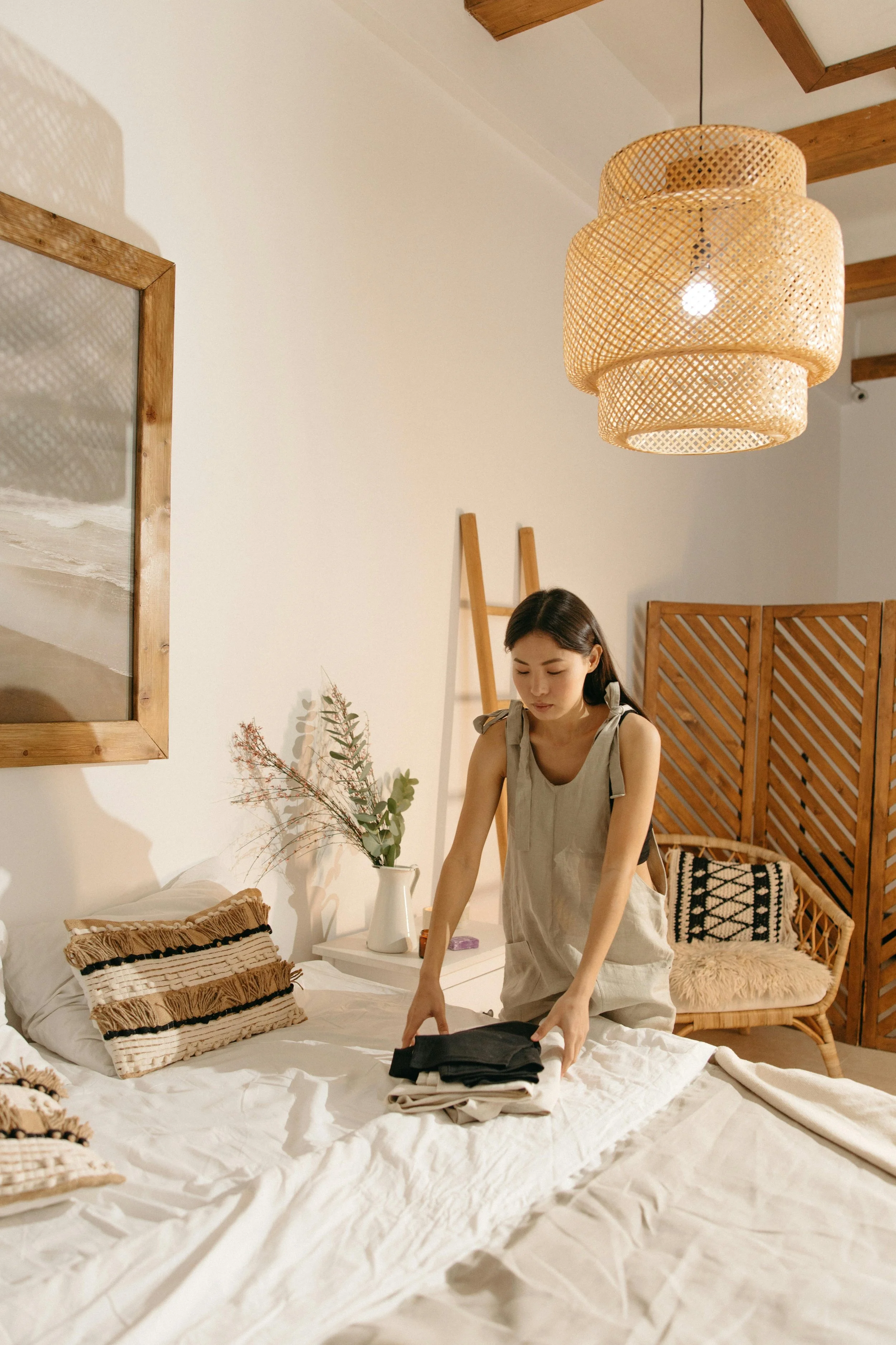 A woman in a beige apron folding clothes on a bed in a bedroom with neutral tones and boho decor.