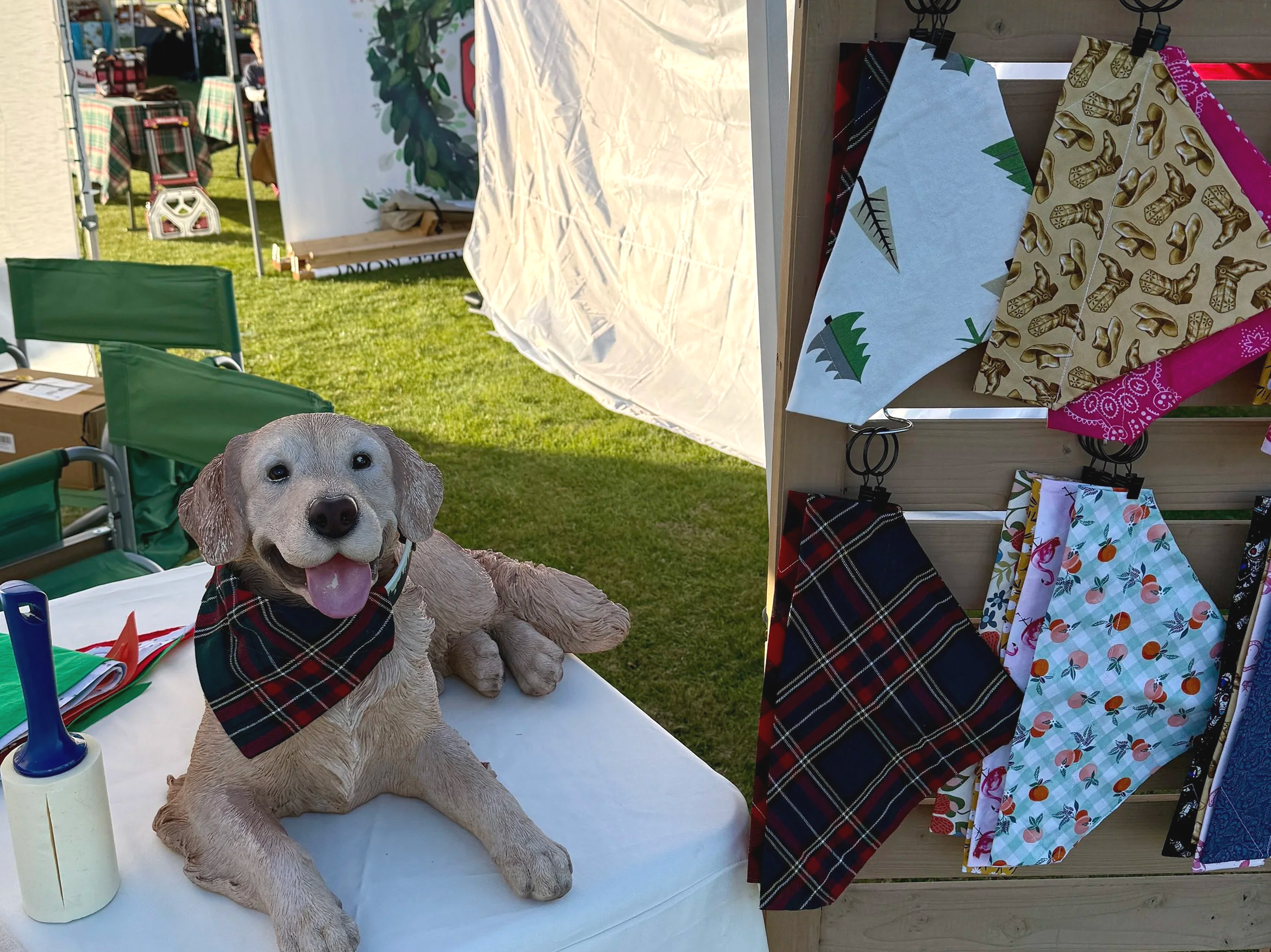 A life-sized dog sculpture with a plaid bandana around its neck, sitting on a table at an outdoor festive event, with a display of patterned cloth napkins hanging on a wooden stand beside it.