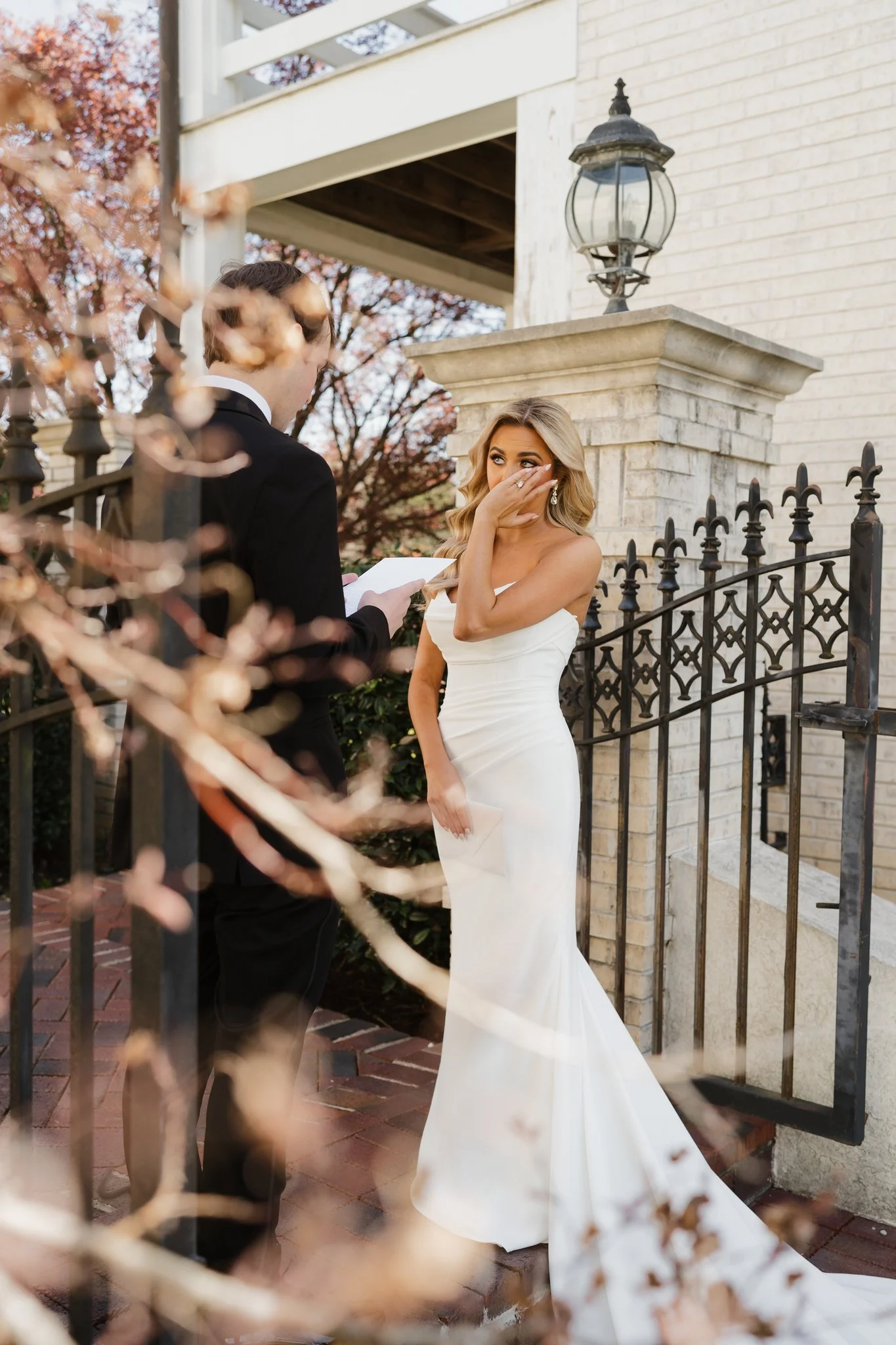A bride in a white wedding gown covers her mouth and looks emotional as she stands outside near a black wrought iron fence, while a man in a black tuxedo reads from a paper during a wedding ceremony in Memphis, TN. 