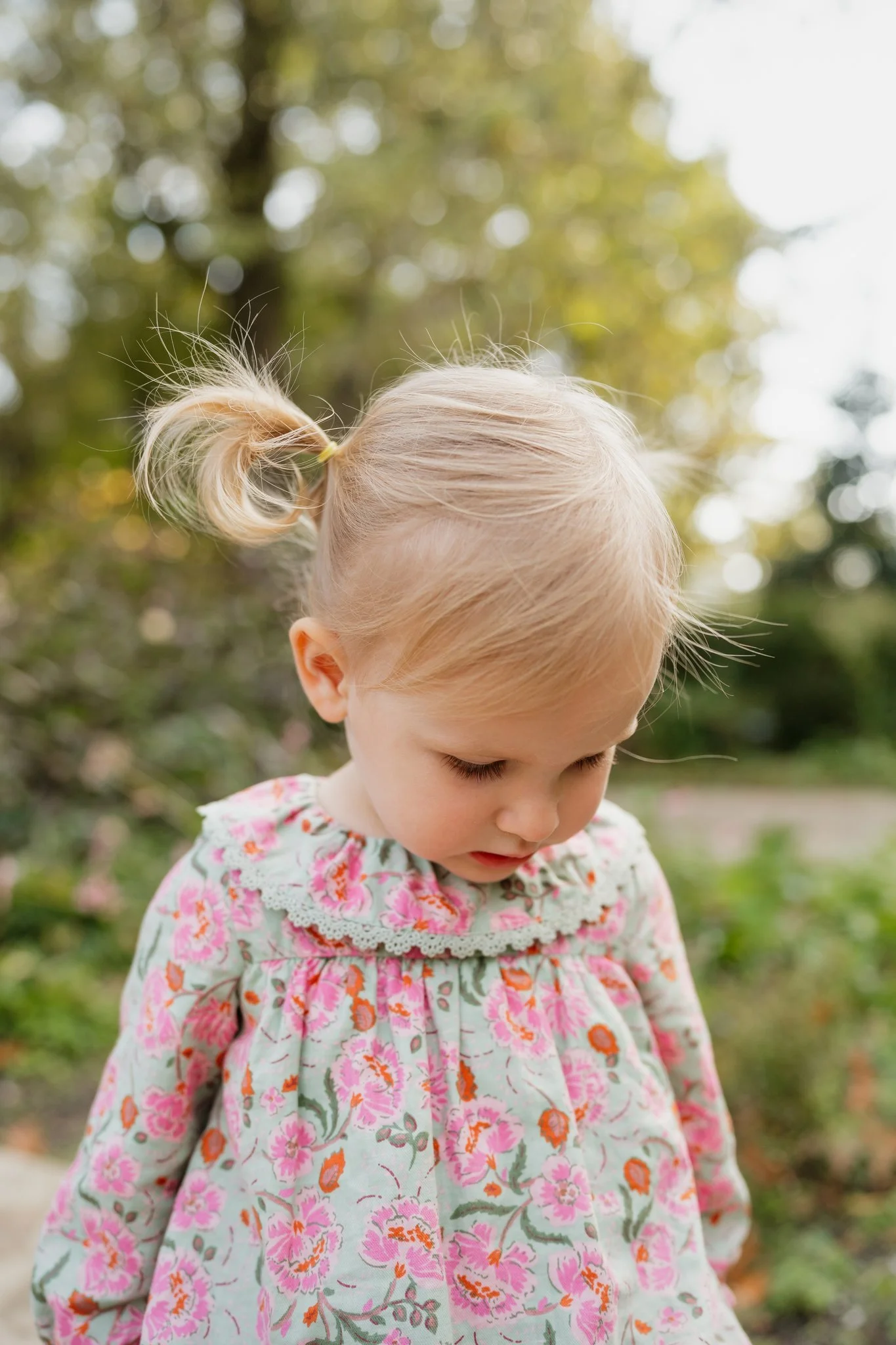 A young girl with blonde hair in a ponytail, wearing a floral dress, looking down outdoors with trees and sunlight in the background in Memphis, TN. 