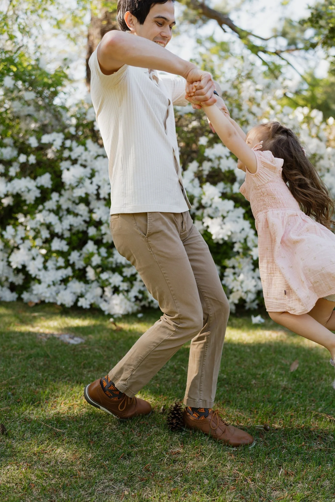 A man and young girl playing outdoors in a garden with blooming bushes, holding hands and spinning around, smiling, dressed casually.