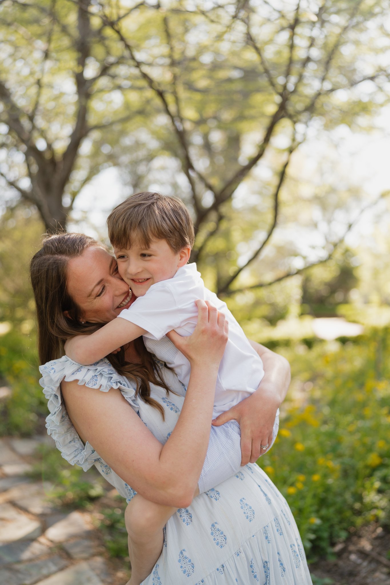 A woman in a white dress with blue embroidery holding a smiling young boy in a white shirt outdoors in a park with green trees and yellow flowers in Memphis, TN. 