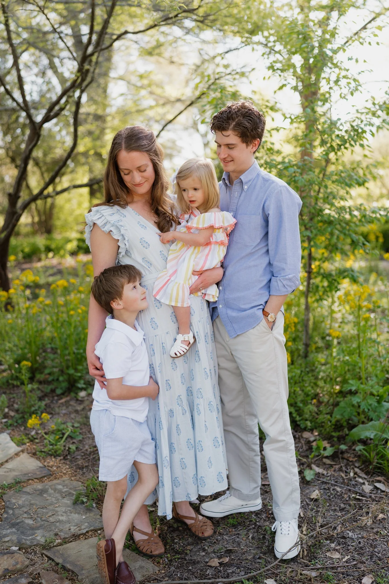 A family of five standing outdoors on a sunny day, surrounded by trees and greenery. The mother holds a young girl, and a boy stands close to her, while the father smiles at the girl. All are dressed in light-colored, casual clothing and appear happy