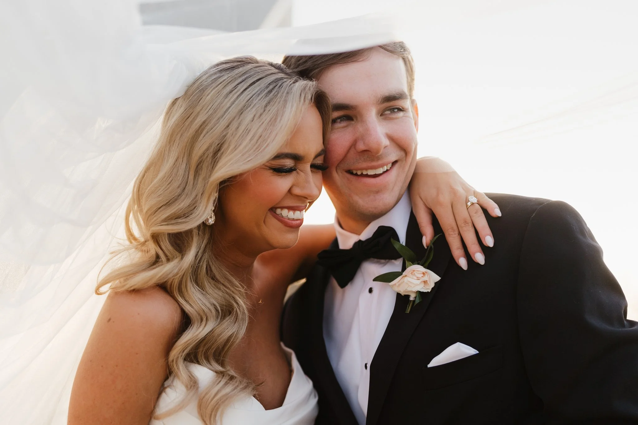 A smiling bride and groom embrace, with the bride's hand on the groom's shoulder, wearing a tuxedo and wedding dress during golden hour at a wedding in Memphis, TN.