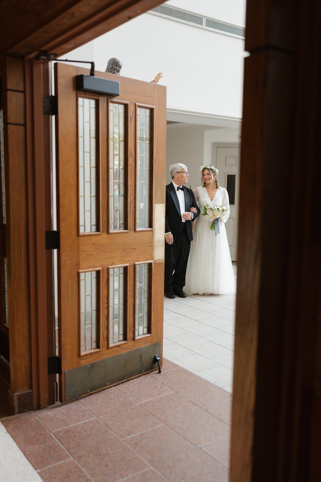 A bride in a white wedding dress holding a bouquet of flowers smiles as she walks with an older man dressed in a black tuxedo and bow tie, possibly her father, in an indoor setting with white walls and tiled floor. The photo is taken through a partia