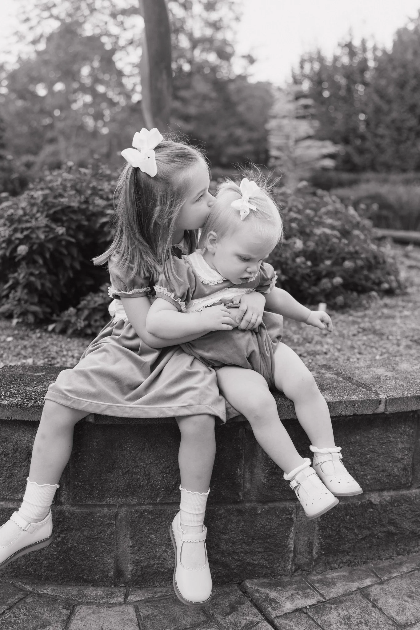 Two young girls sitting on a stone ledge outdoors, with the older girl kissing the younger girl on the head. Both are wearing dresses, socks, and shoes, and have flowers in their hair. The background includes trees and a light sky.