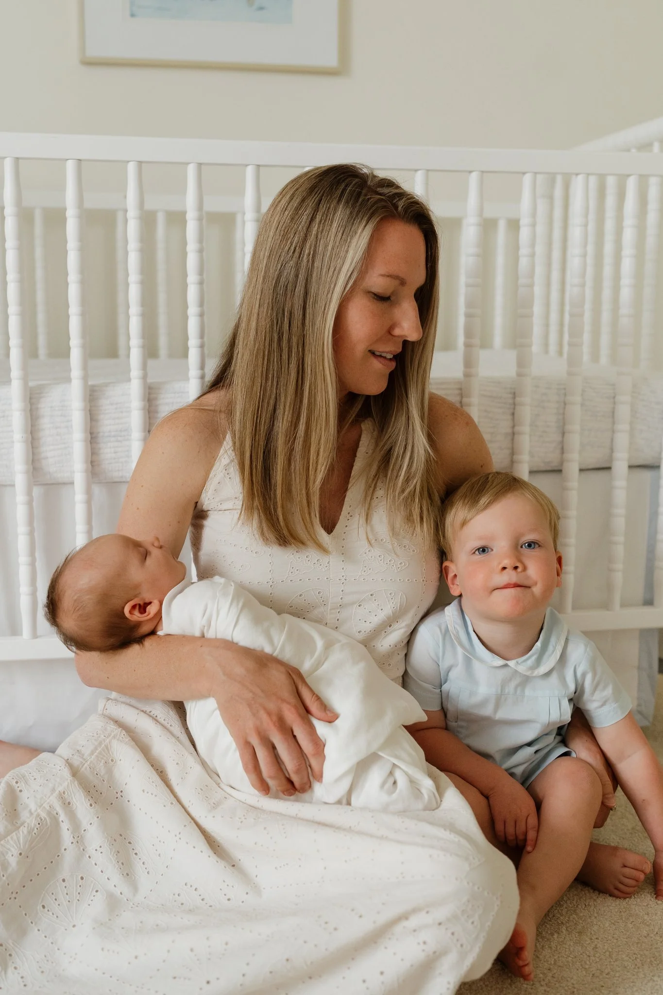 A woman with long blonde hair holding two children, one in each arm, while sitting on the floor in a room with white walls and a white crib in the background. The girl in the foreground has blond hair and blue eyes, and the boy next to her has light 