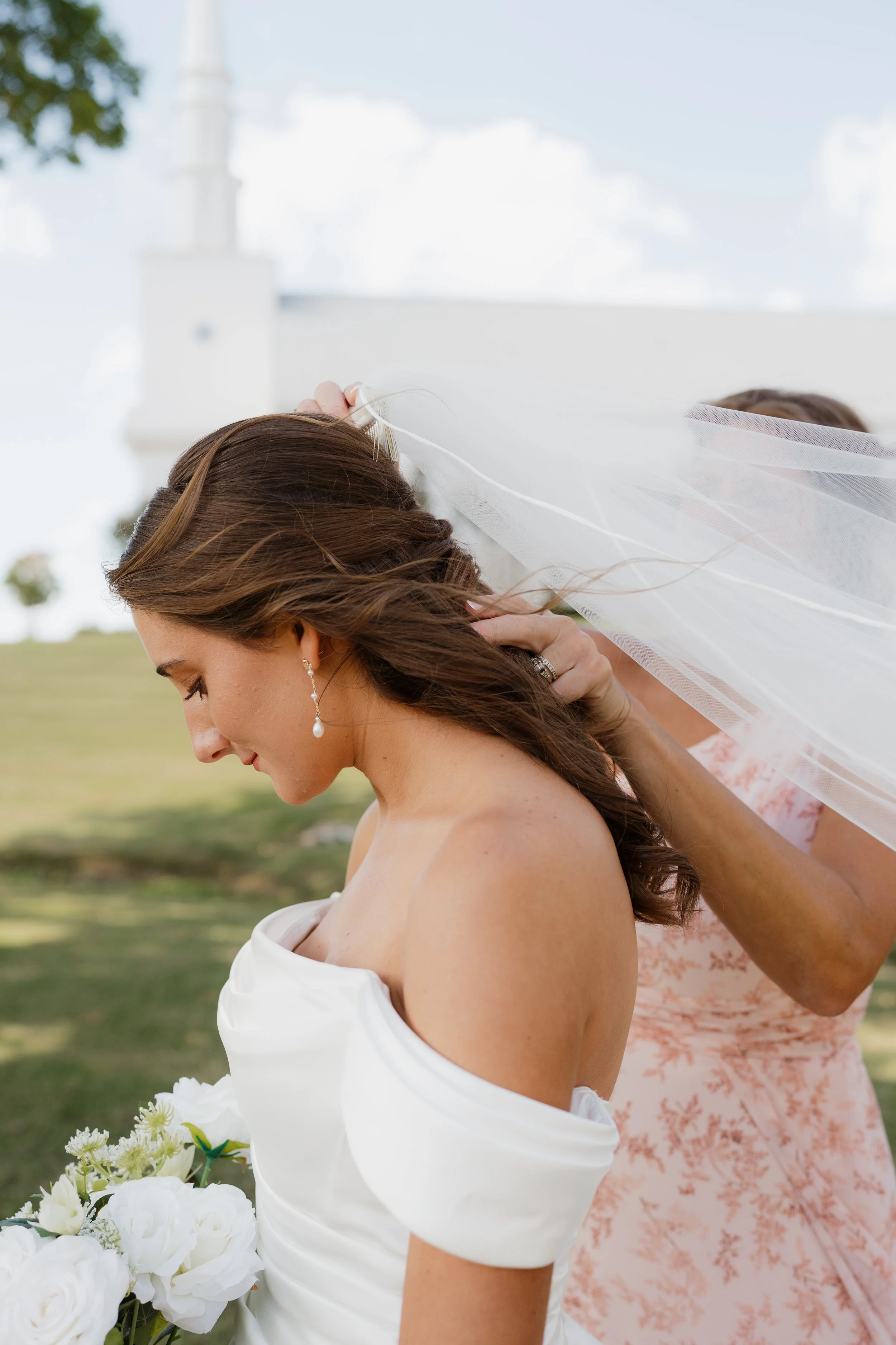 A bride with long brown hair and pearl earrings, wearing a white off-shoulder wedding dress, holding a bouquet of white flowers, with another woman in pink floral dress adjusting her veil outdoors in Memphis, TN. 