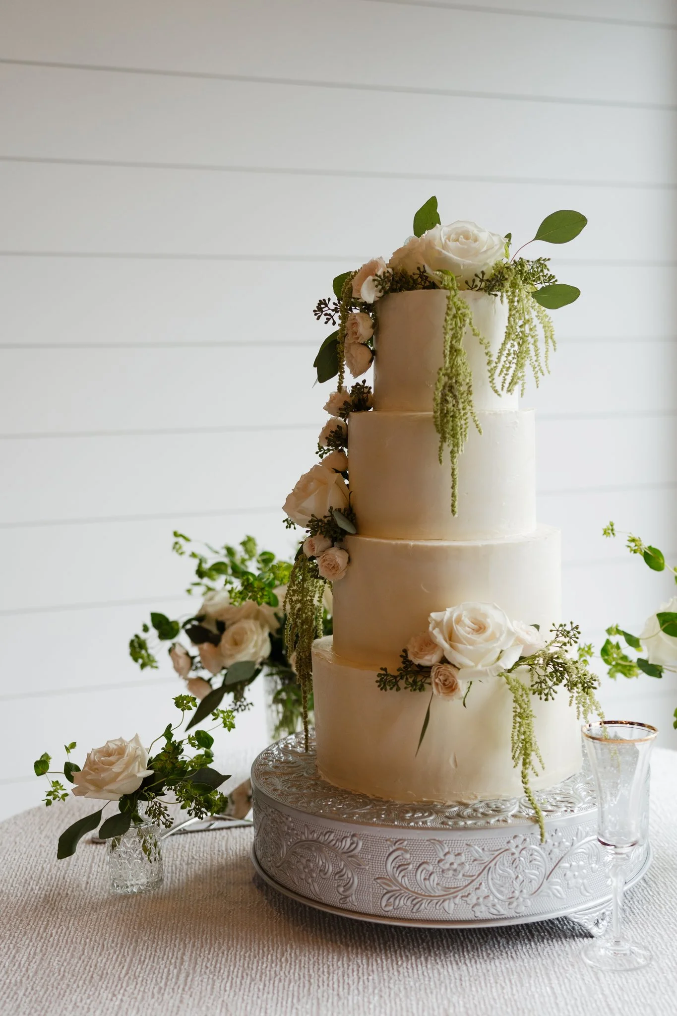 A four-tiered white wedding cake decorated with white roses, greenery, and hanging green amaranthus, placed on an ornate silver cake stand on a textured tablecloth. Additional flowers and greenery are arranged around the base of the cake in Memphis, 