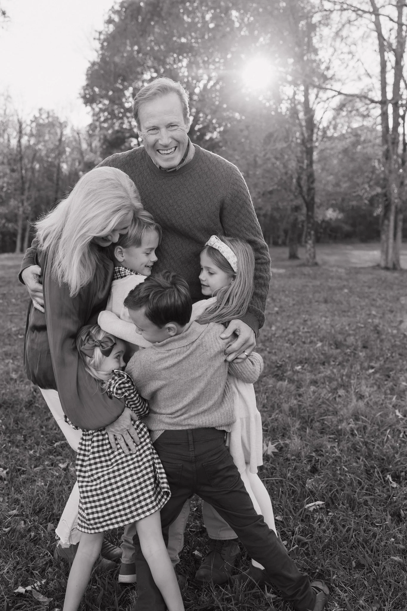 A group of children and an adult are hugging outdoors on a grassy field, with trees and sunlight in the background, in a joyful moment in Memphis, TN. 