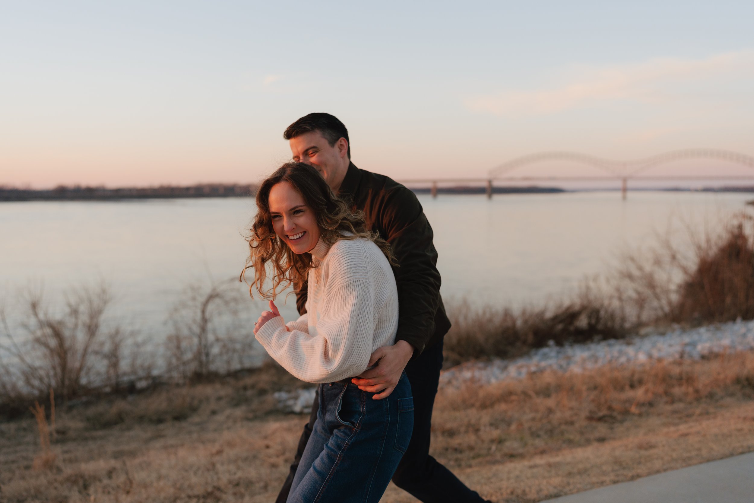 A young couple is outdoors near a body of water, with a bridge in the background, during sunset. The man is hugging the woman from behind, and both are smiling and appear happy in Memphis, TN. 