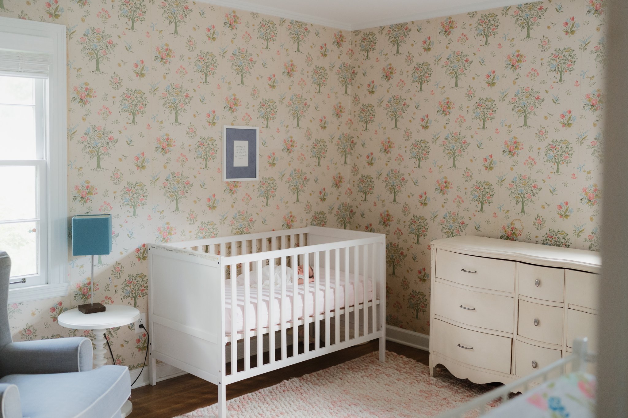 A nursery room with floral wallpaper, a white crib with a sleeping baby, a blue table lamp on a round white table, a window, and a white dresser in Memphis, TN. 