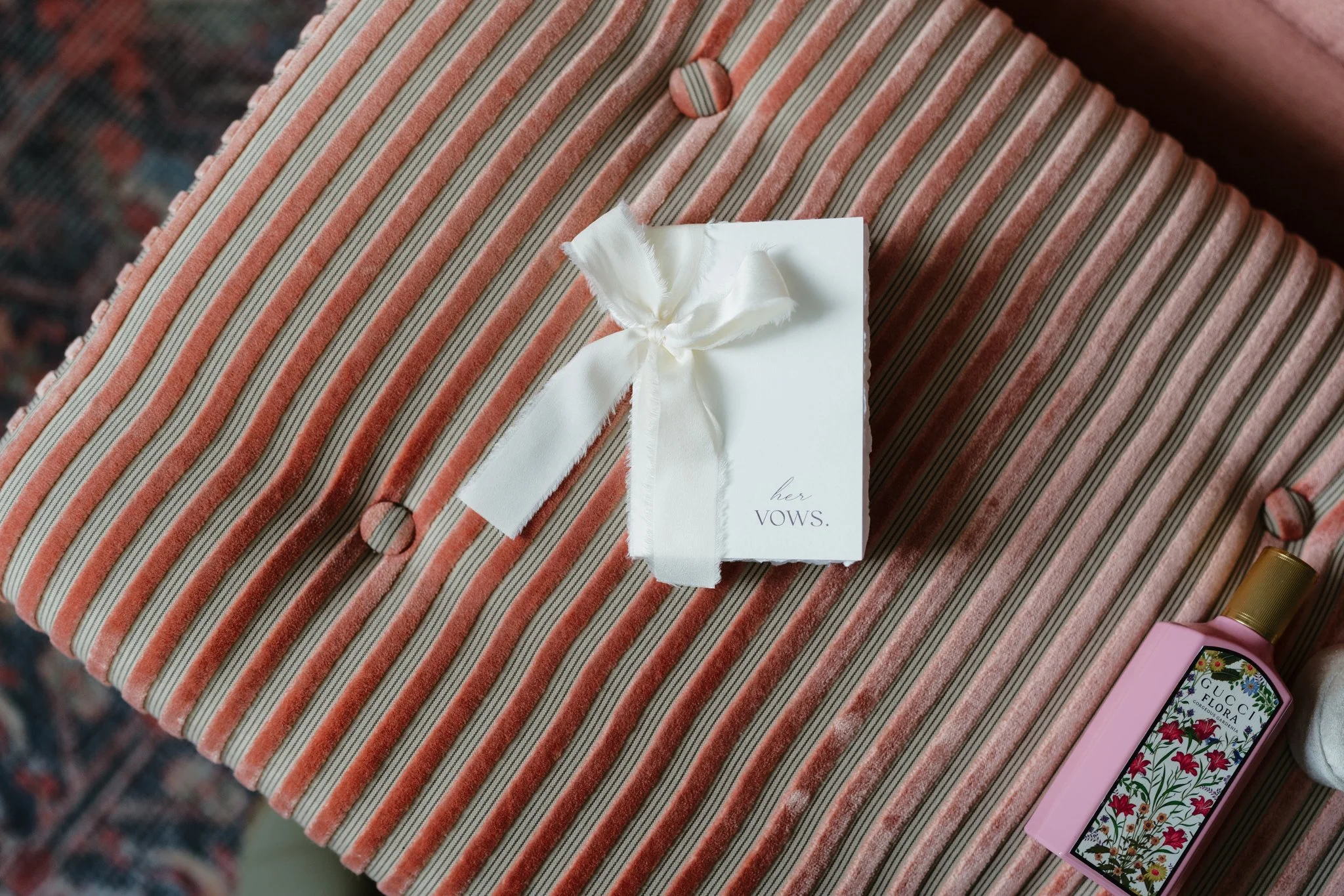 Close-up of a striped fabric with a white ribbon and a small white card that reads 'her VOWS' on top of it. There are a few small items near the fabric, including a tube of pink lipstick and a floral-patterned packaging.