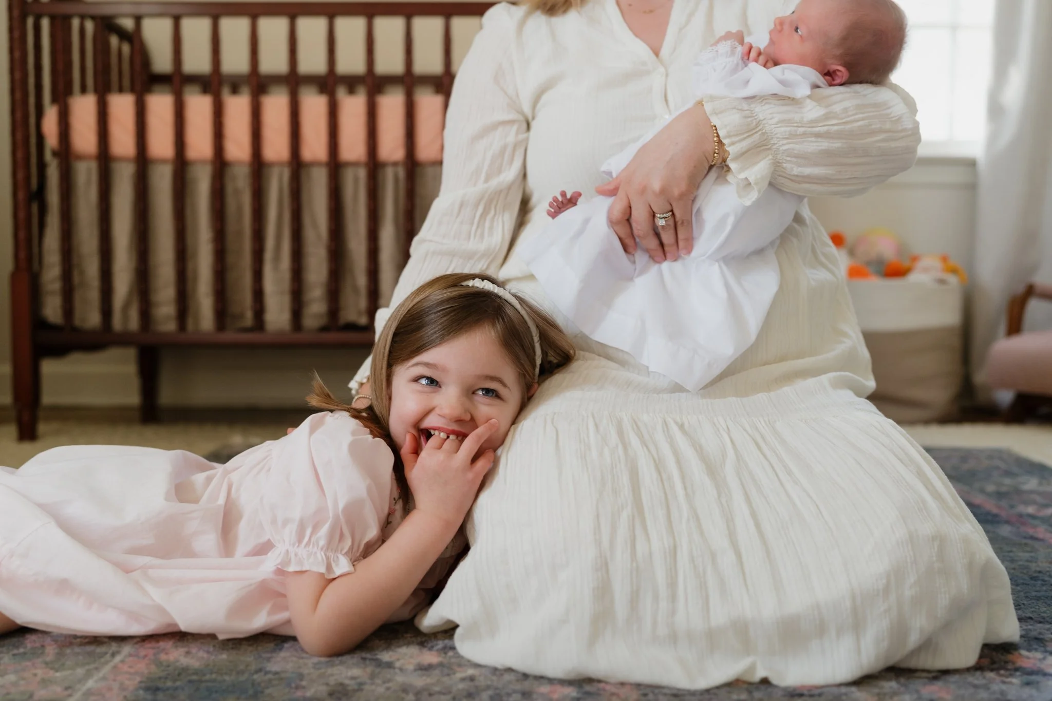 A young girl lying on a carpeted floor smiling, with her hand near her mouth, while an adult holding a baby is in the background. The baby is looking at the adult and is dressed in white. The adult is wearing a cream-colored dress or blouse in Memphi