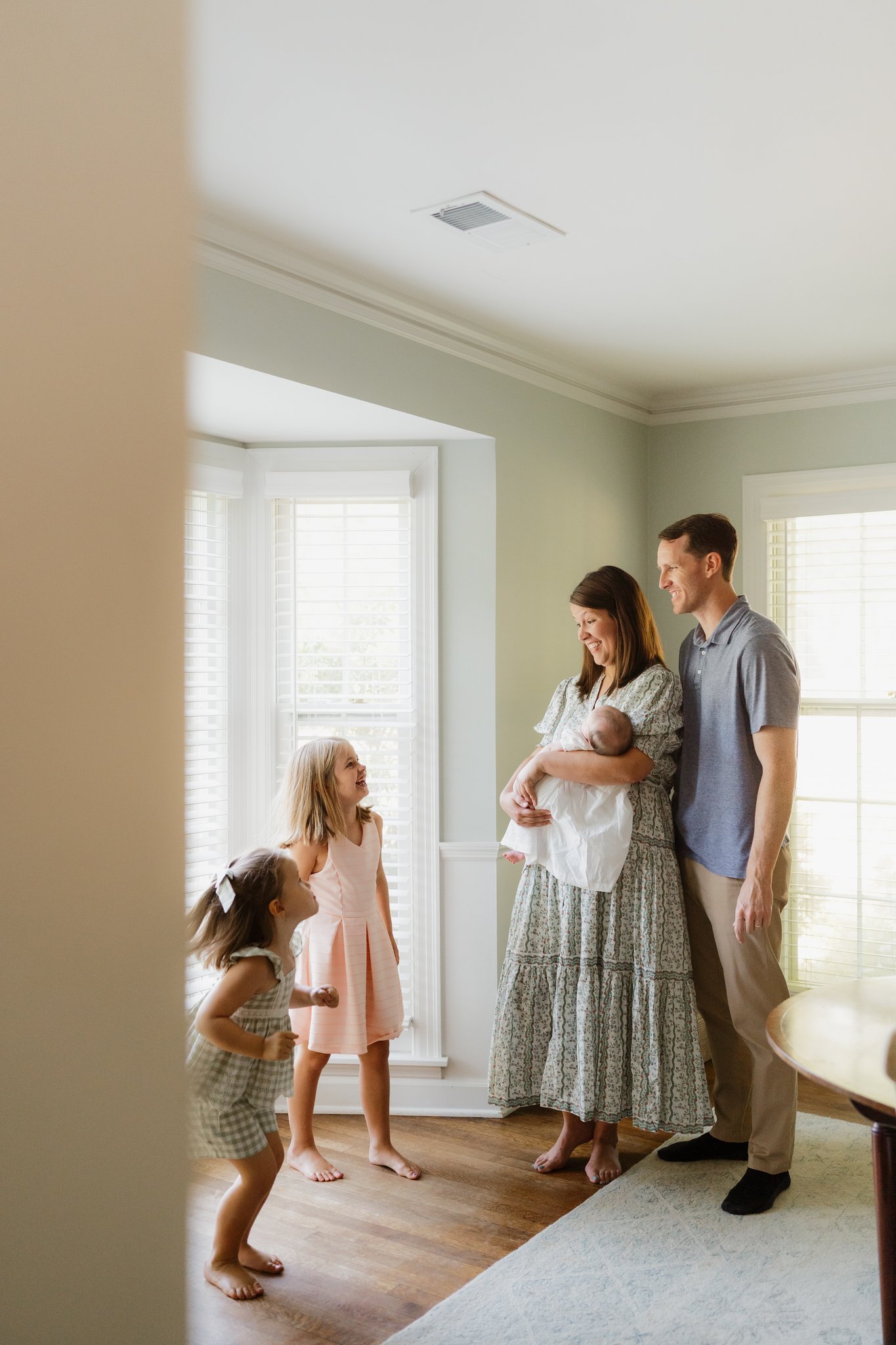A family of five, including two young girls, a woman, and a man, gathered in a bright, welcoming room with large windows and white shutters. They appear to be joyfully interacting, possibly during a family moment or visit in Memphis, TN. 