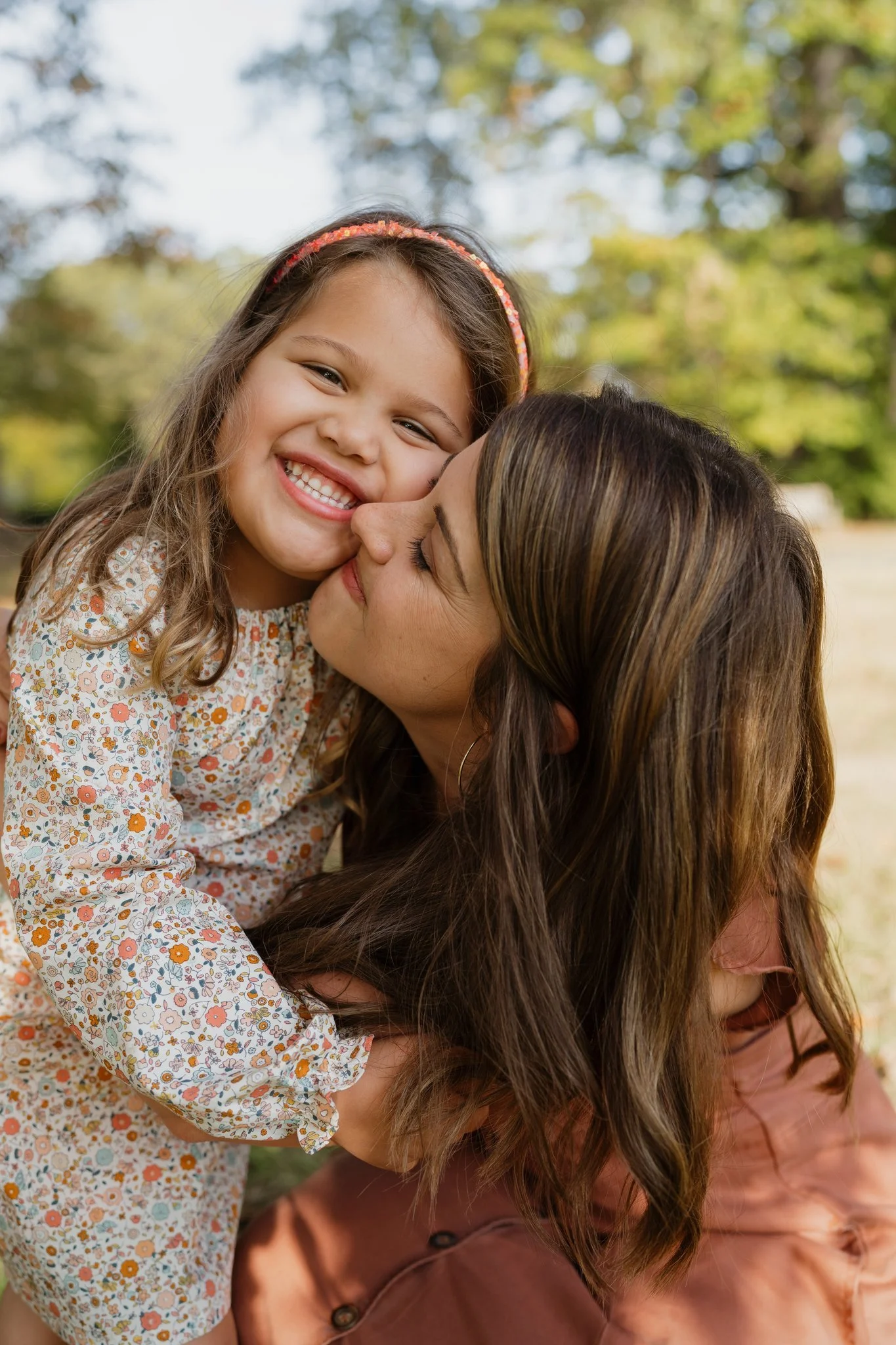 A smiling young girl with light brown hair wearing a floral dress is being hugged by a woman with long brown hair. They are outdoors with trees and sunlight in the background.