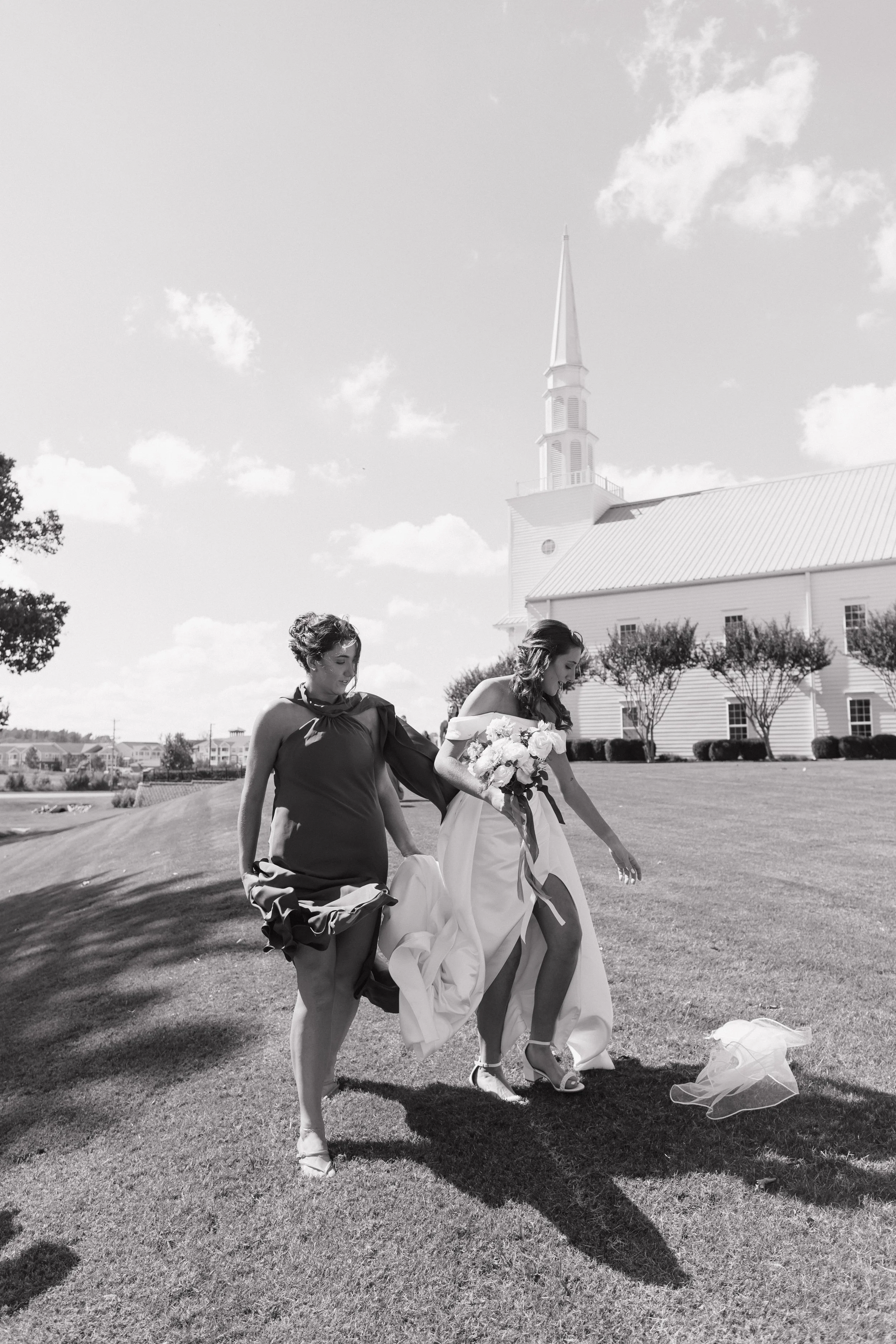 A bride in a wedding dress and heels walking with two women on a lawn outside a church with a tall steeple and a white exterior. One woman wears a dark dress, and the other is in a dark top and a skirt, holding the bride's hand in Memphis, TN. 