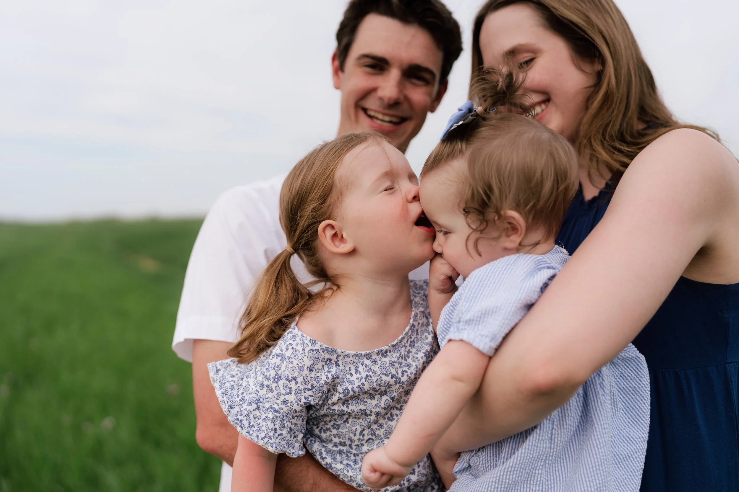 A family of four, including a man, a woman, and two young girls, hugging each other outdoors on a grassy field with cloudy sky in the background, smiling and showing affection in Memphis, TN. 
