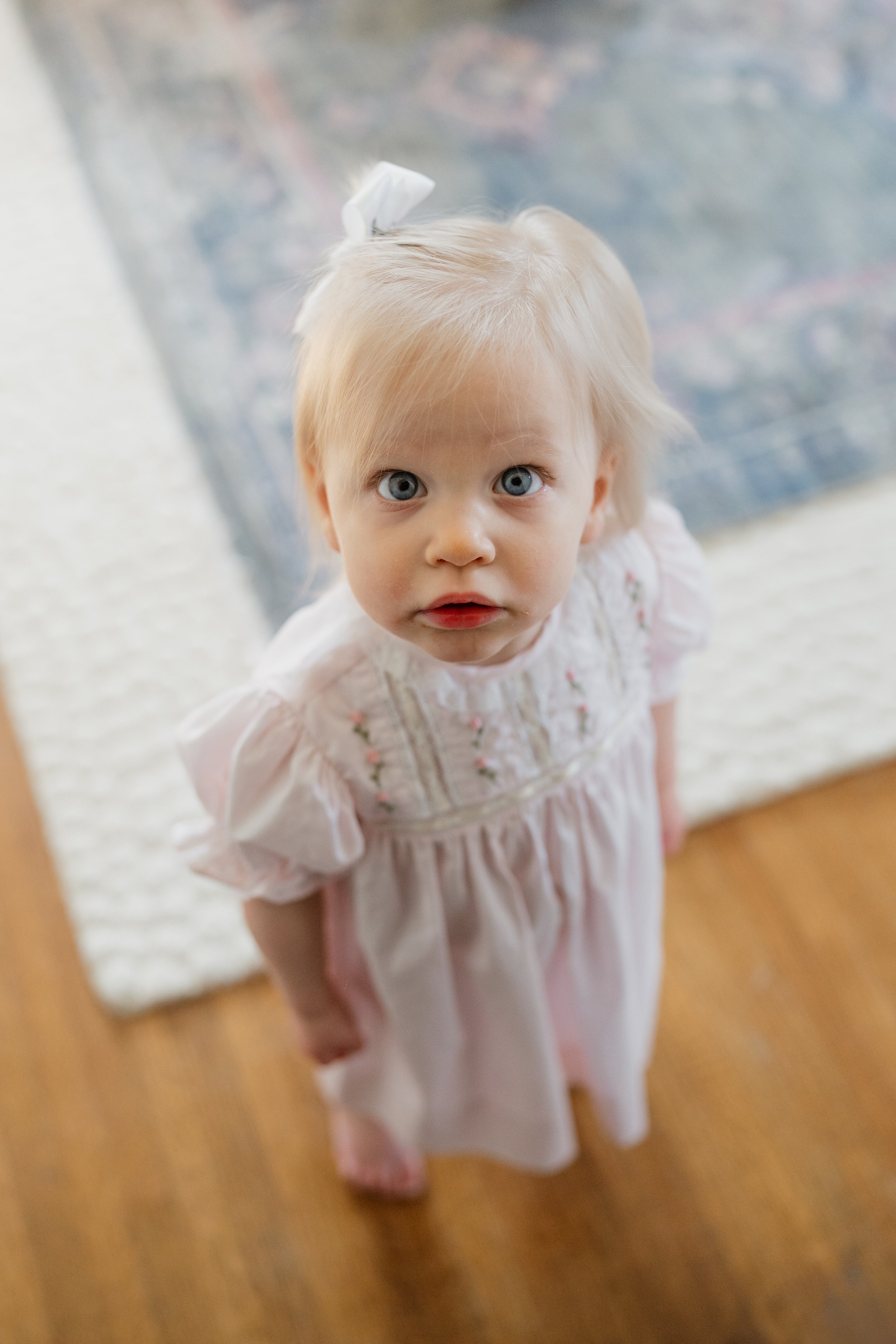 A young girl with blonde hair and blue eyes looking up at the camera through a mirror. She is wearing a light pink dress with floral embroidery in Memphis, TN. 