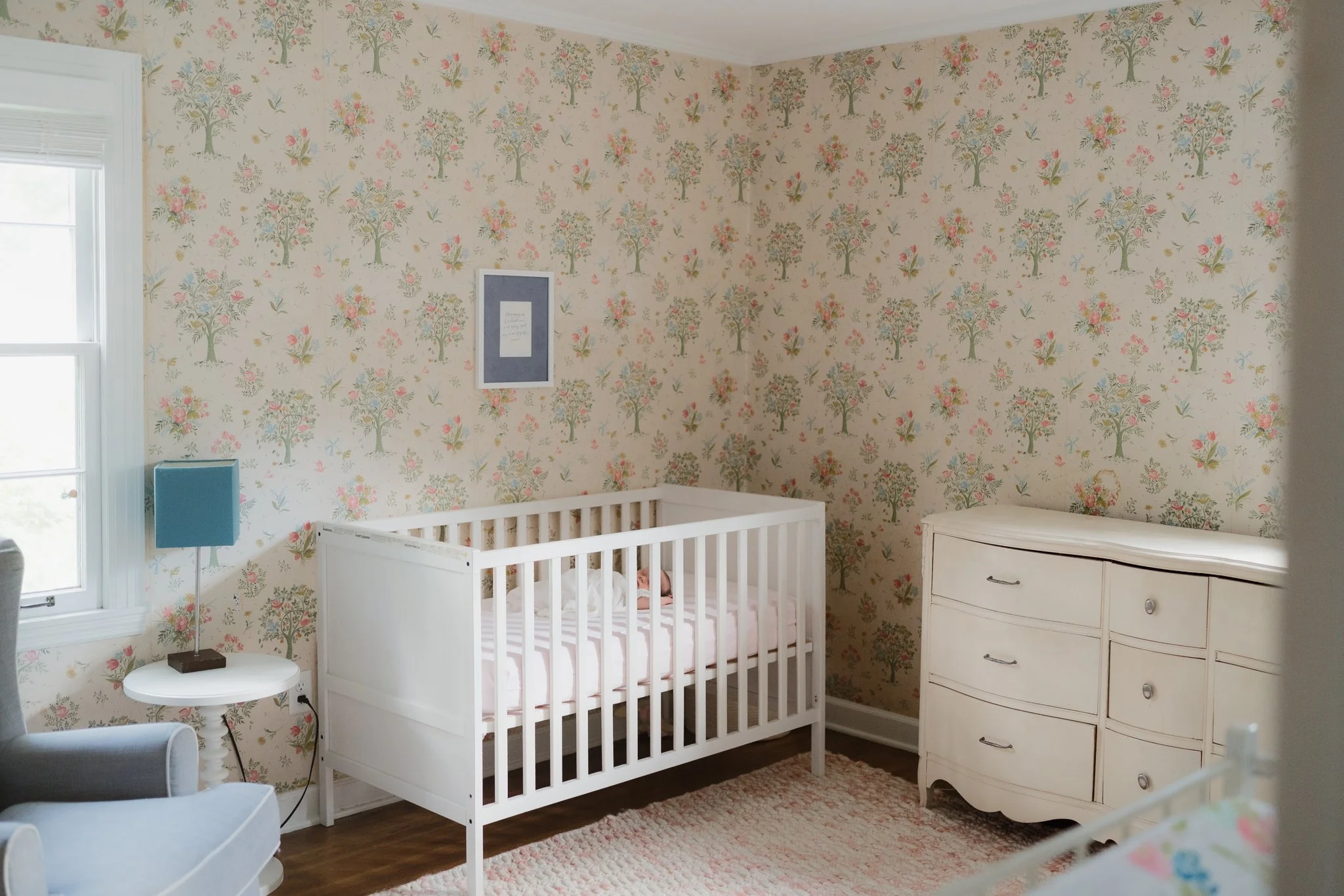 A nursery room with floral wallpaper, a white crib, a small blue table lamp, a white dresser, a window with white blinds, and a stuffed animal inside the crib in Memphis, TN. 