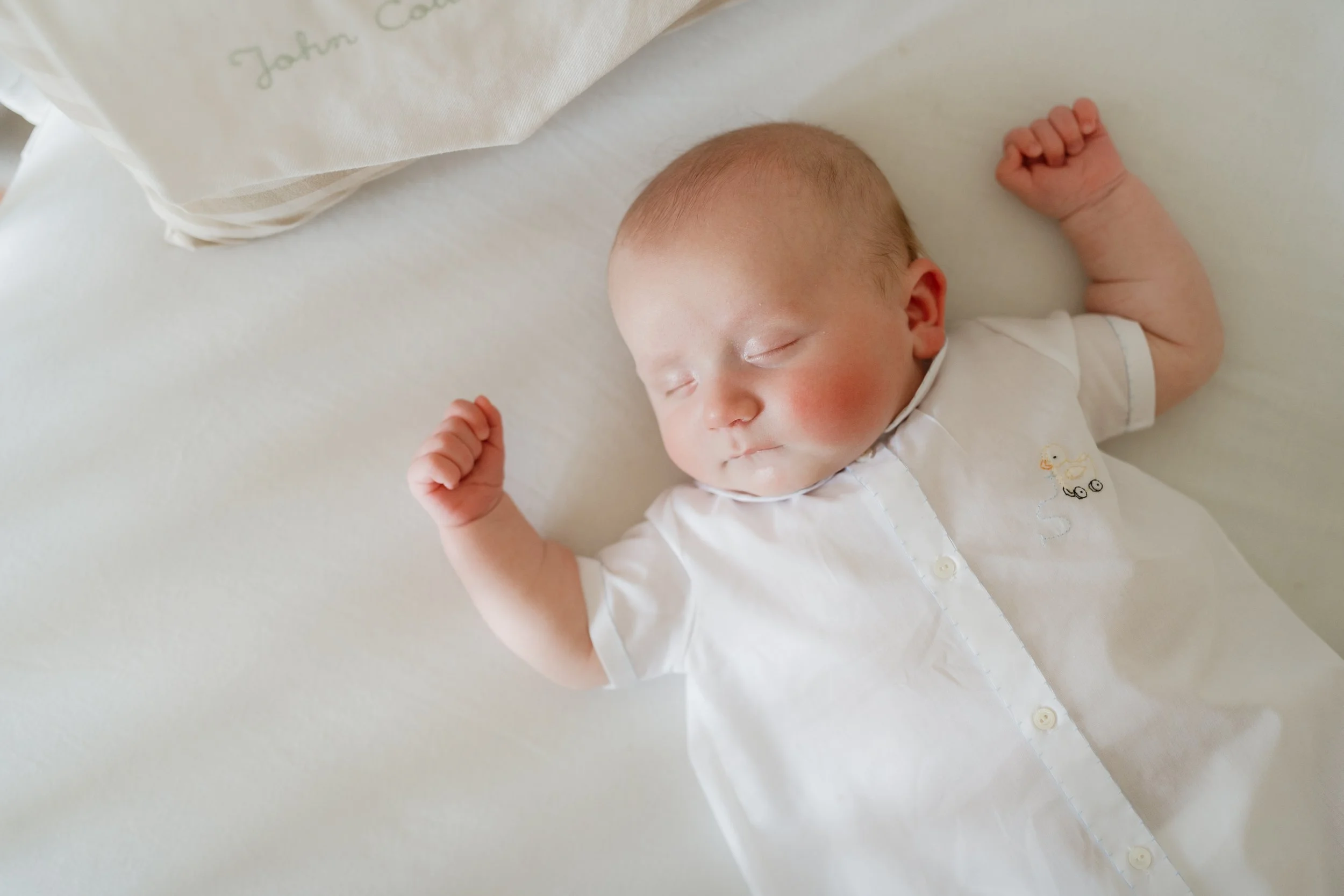 A sleeping baby lying on a white bed with a white blanket beside him, wearing a white outfit with a duck embroidery in Memphis, TN. 