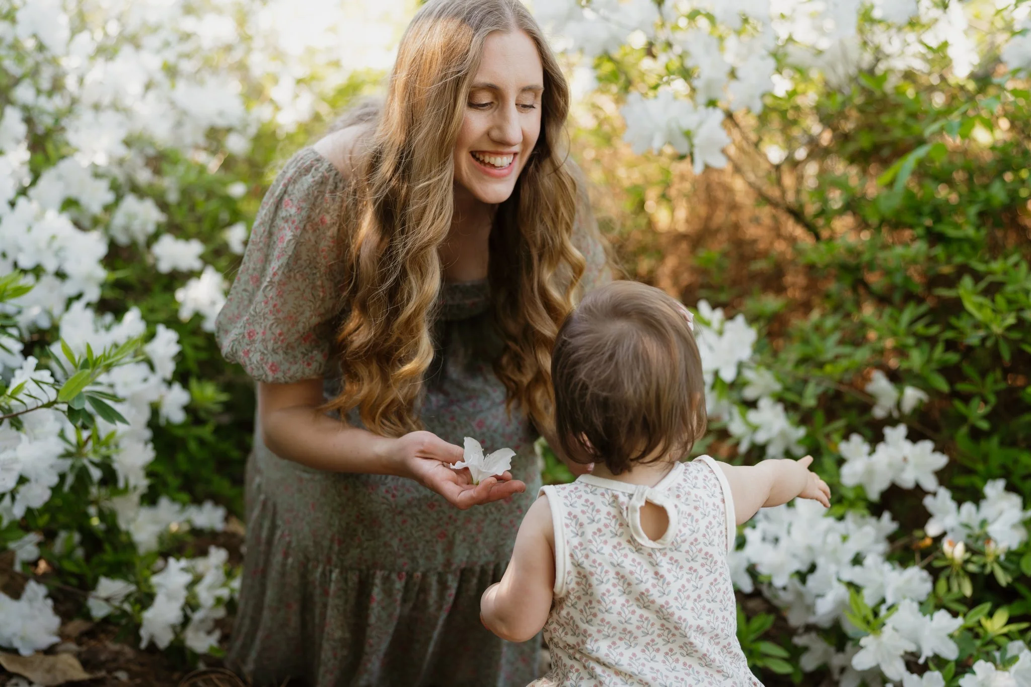 A woman with long wavy hair smiling as she watches a young girl pointing at white flowers in a garden in Memphis, TN. 