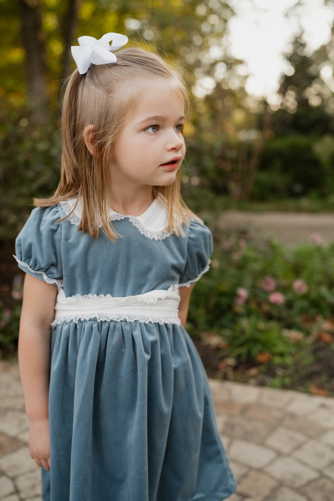 A young girl with blonde hair wearing a blue dress with lace details and a white bow in her hair, standing outdoors with blurred trees and pink flowers in the background.