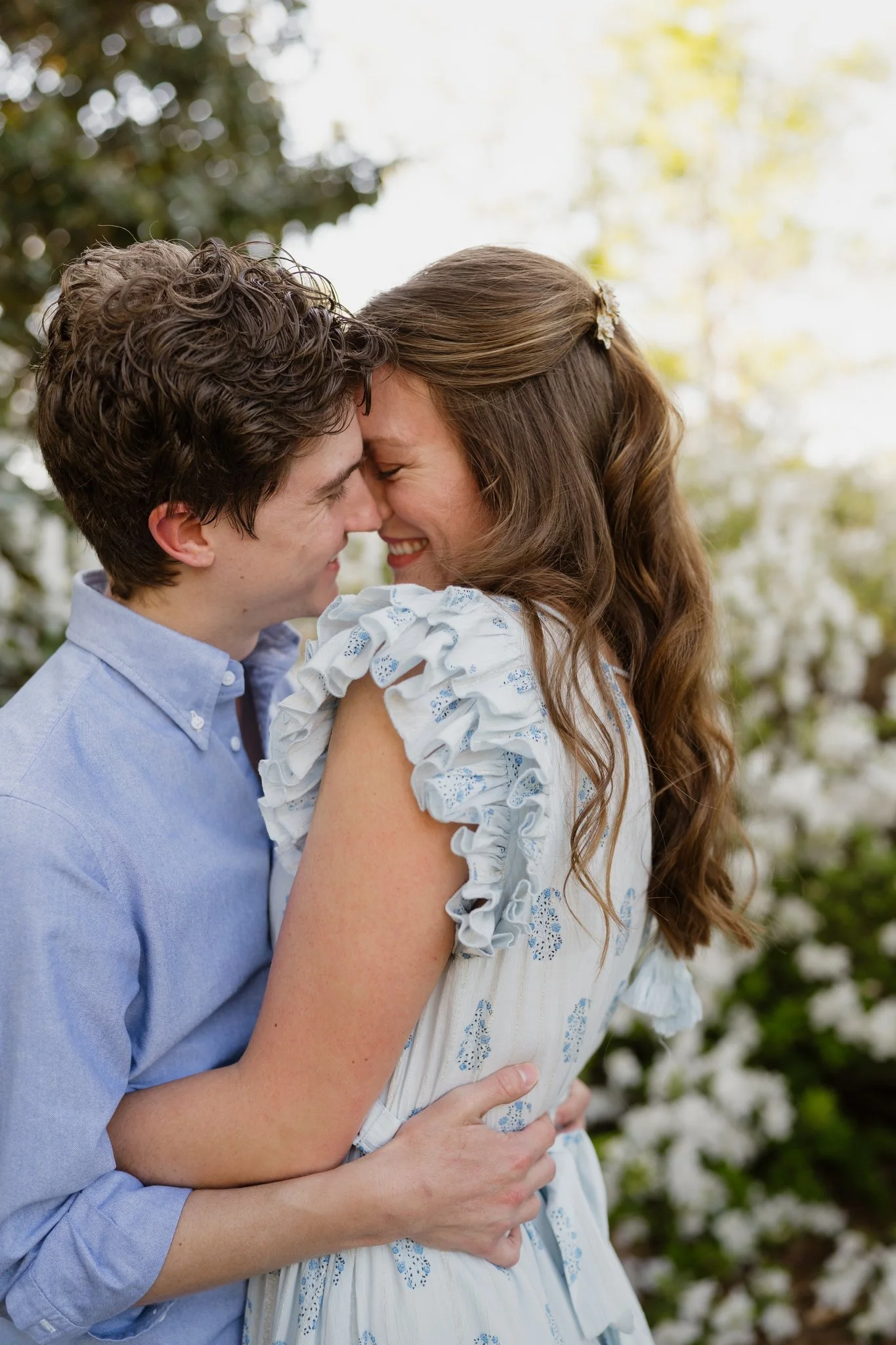 A couple standing outside, holding each other closely and touching foreheads, smiling with their eyes closed. The man has curly hair and is wearing a light blue shirt, and the woman has long wavy hair with a hair clip, wearing a white dress with blue