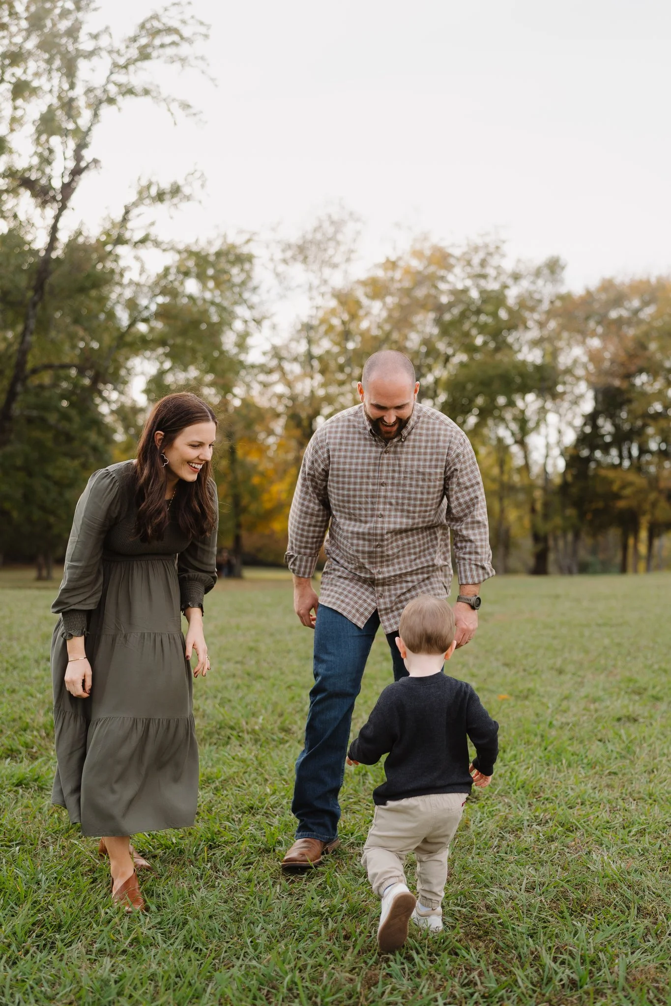 A family of four enjoying a day outdoors in a park during fall. The mother and father are talking to their young son who is walking towards them, with the mother smiling and wearing a long green dress, and the father smiling and wearing a plaid shirt