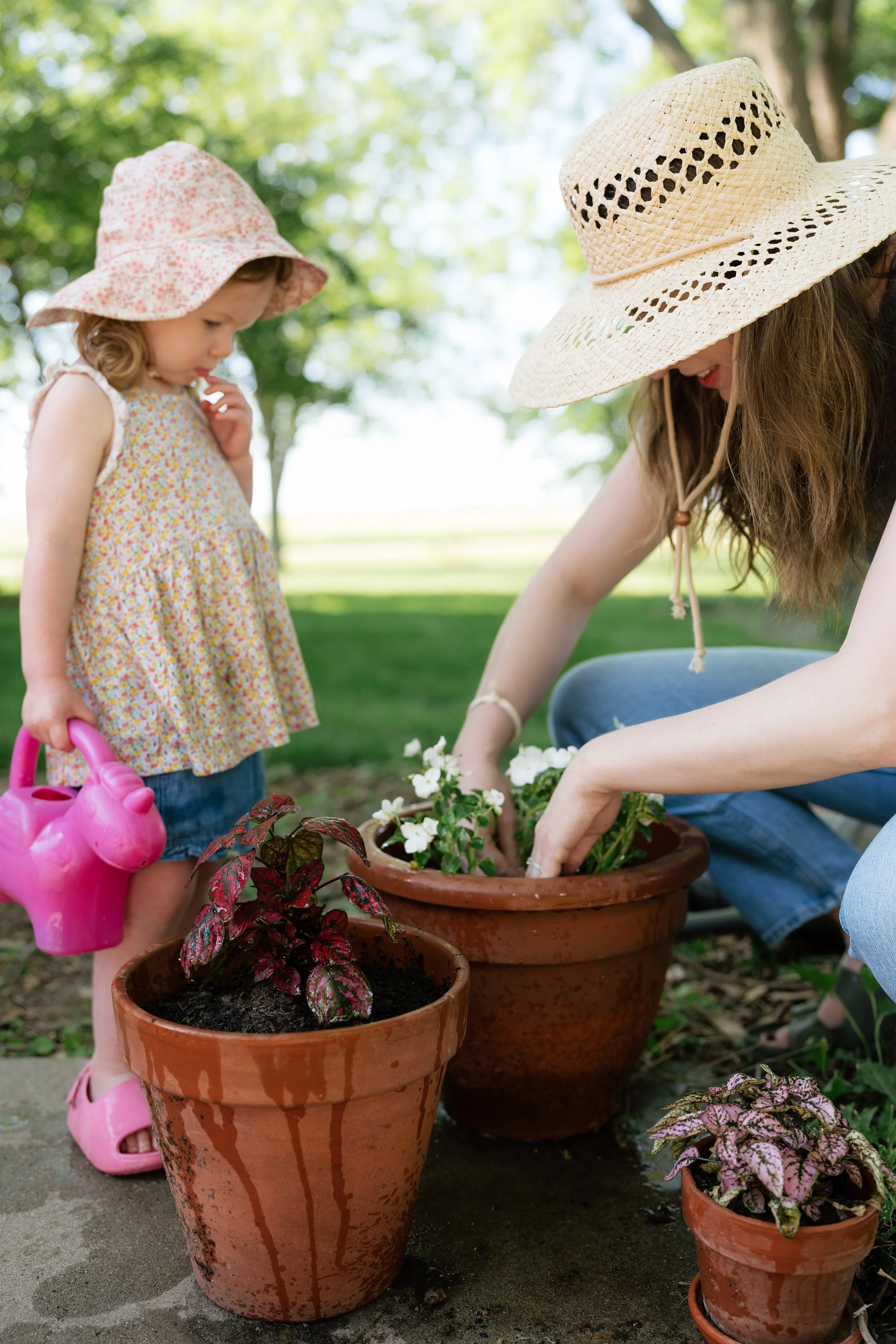 A woman and a young girl planting flowers in outdoor pots. The girl holds a pink watering can and wears a sun hat, while the woman wears a straw hat and is planting flowers in a terracotta pot. There are multiple flowerpots with plants, and trees and
