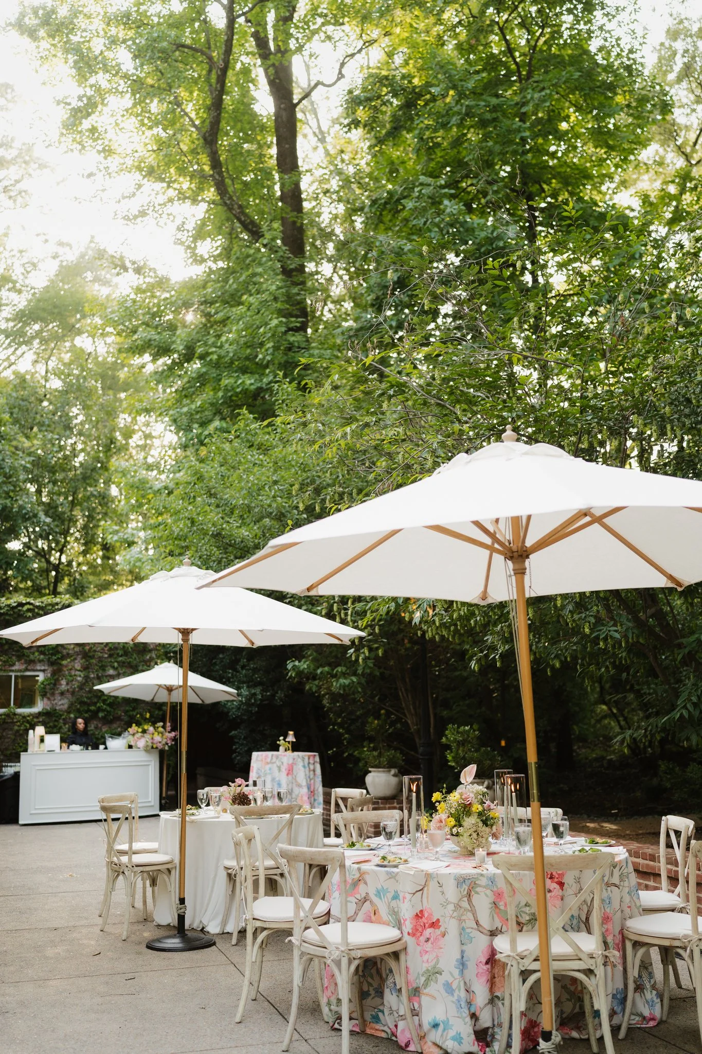 Outdoor event setup with tables decorated with floral tablecloths, floral centerpieces, and white chairs, surrounded by greenery with large white umbrellas providing shade in Memphis, TN. 